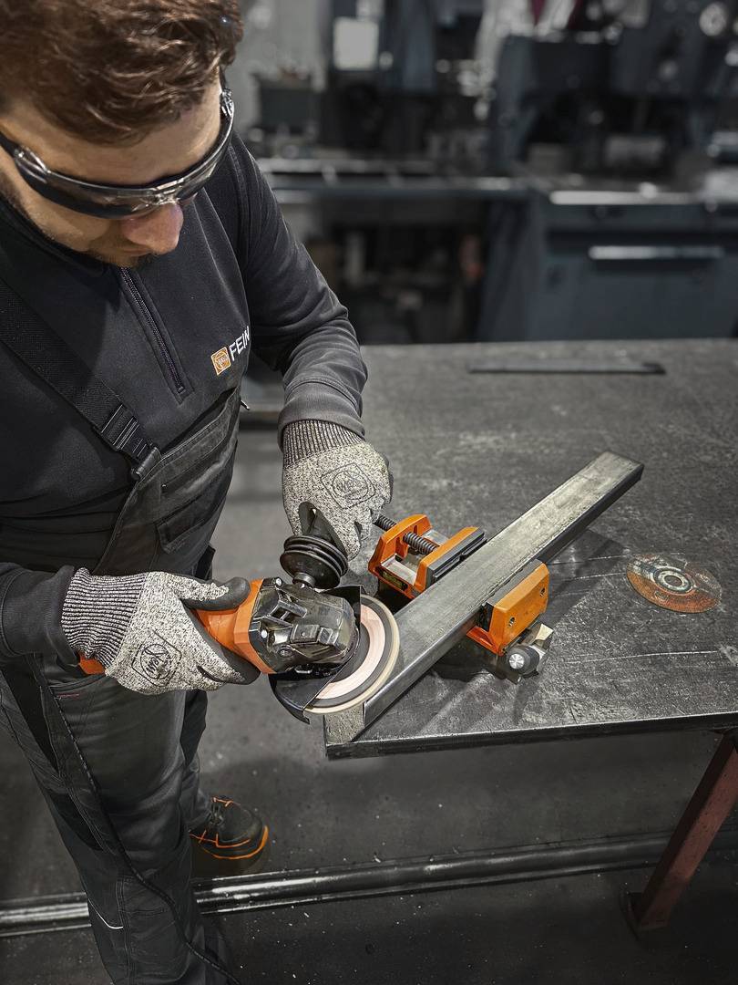 Eine Person bearbeitet mit einem Winkelschleifer ein Metallstück, das in einem Schraubstock auf einem Werktisch befestigt ist.