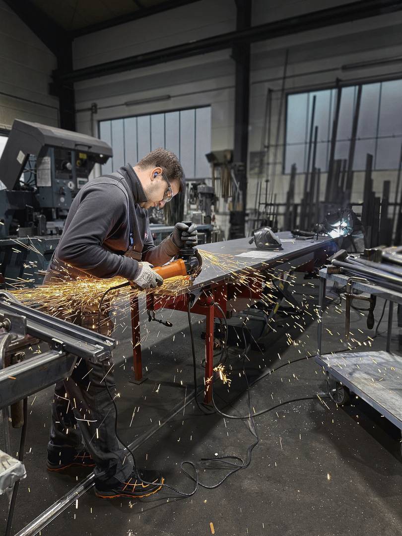 Ein Arbeiter trägt Gehörschutz und schleift Metall in einer Werkstatt. Funken fliegen von der Schleifmaschine. Im Hintergrund sind Industriemaschinen sichtbar.