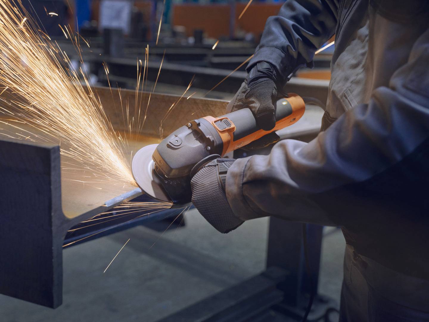 Eine Person schleift Metall mit einem Winkelschleifer, während Funken sprühen. Sie trägt Handschuhe und Arbeitskleidung in einer Werkstatt.