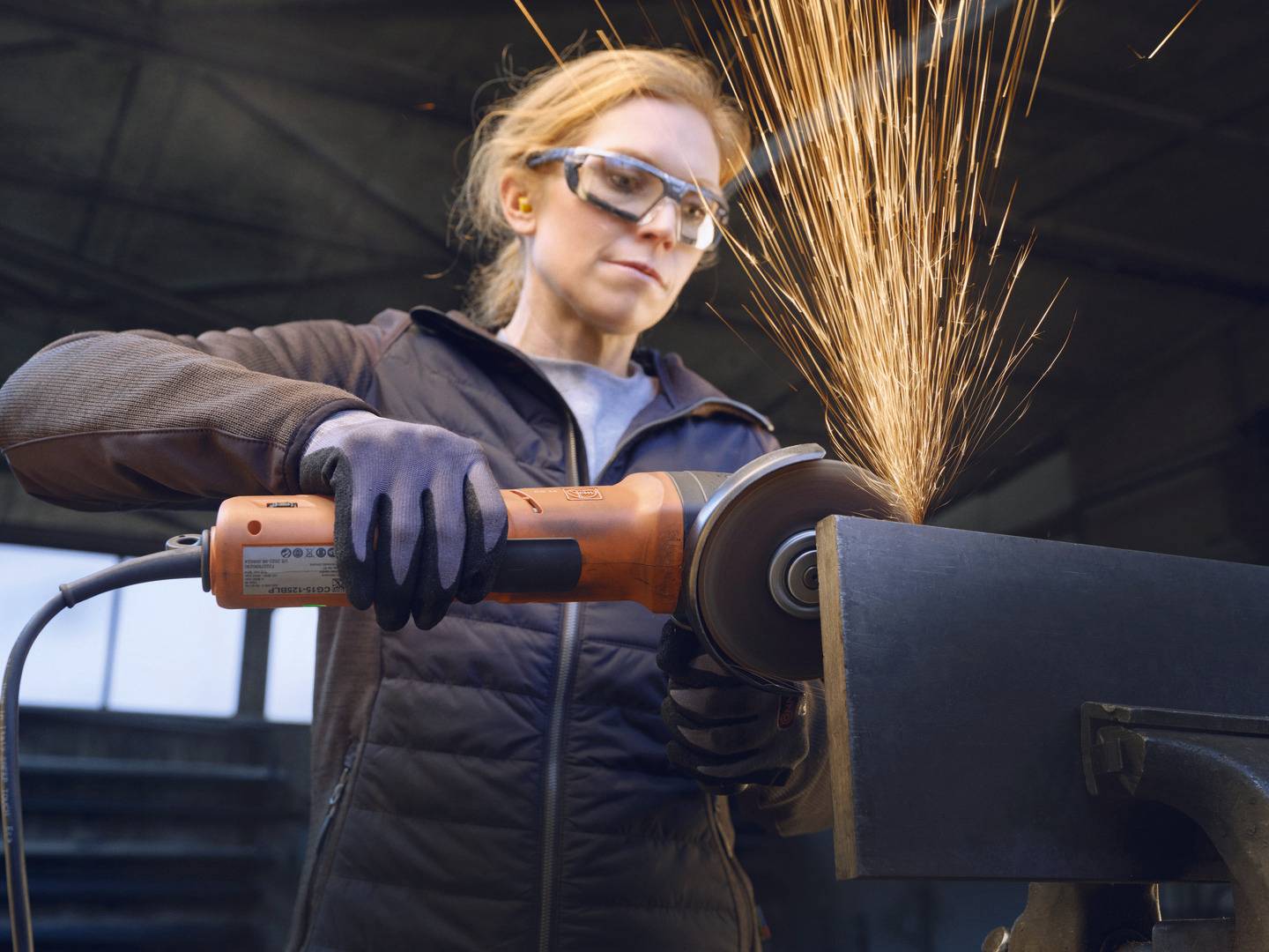 Eine Frau schleift Metall mit einem Winkelschleifer, Funken fliegen. Sie trägt Sicherheitsbrille und Handschuhe in einer Werkstatt.