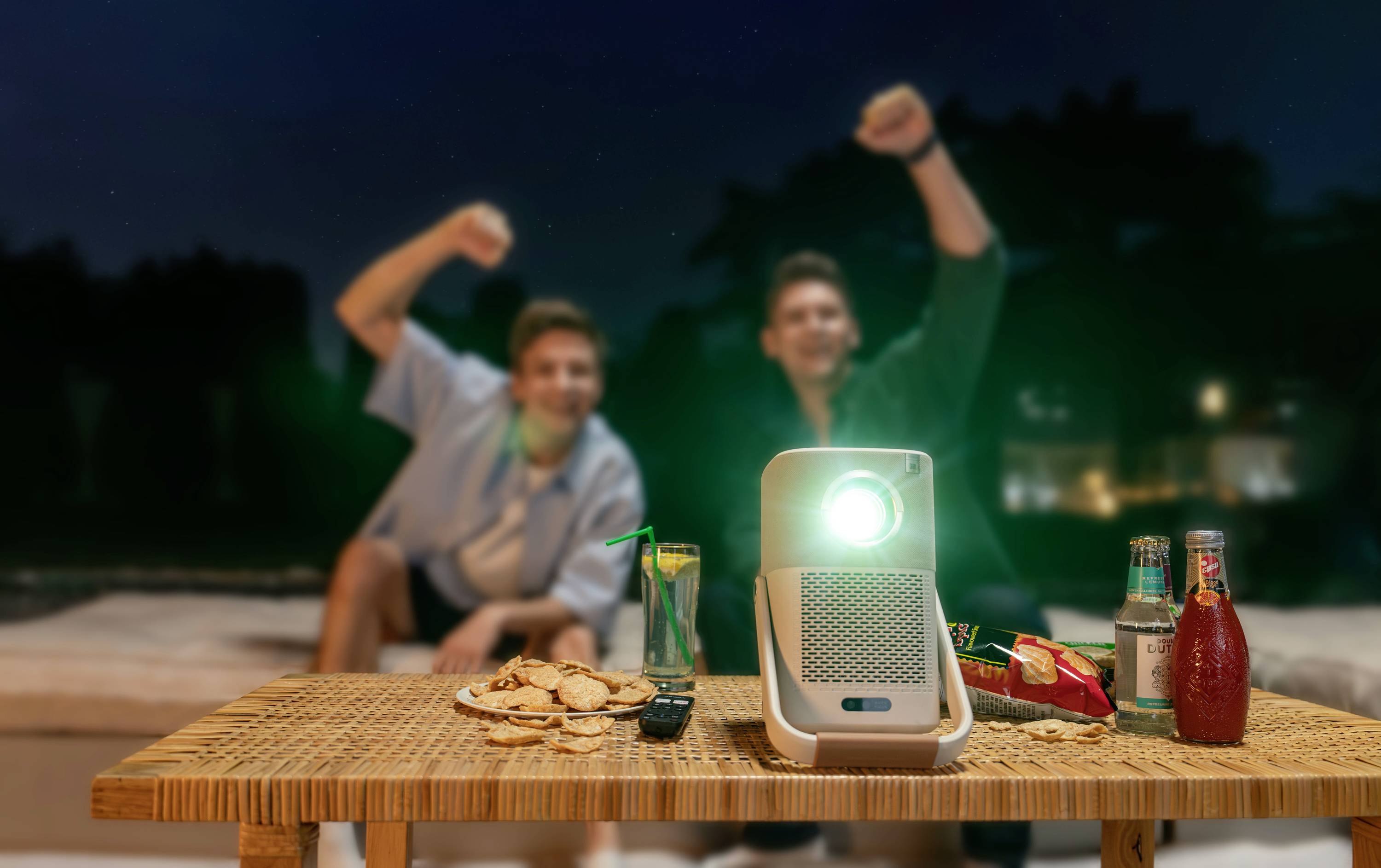 Zwei Personen jubeln auf einem Sofa im Freien, während auf einem Tisch ein Projektor leuchtet, umgeben von Getränken und Snacks.