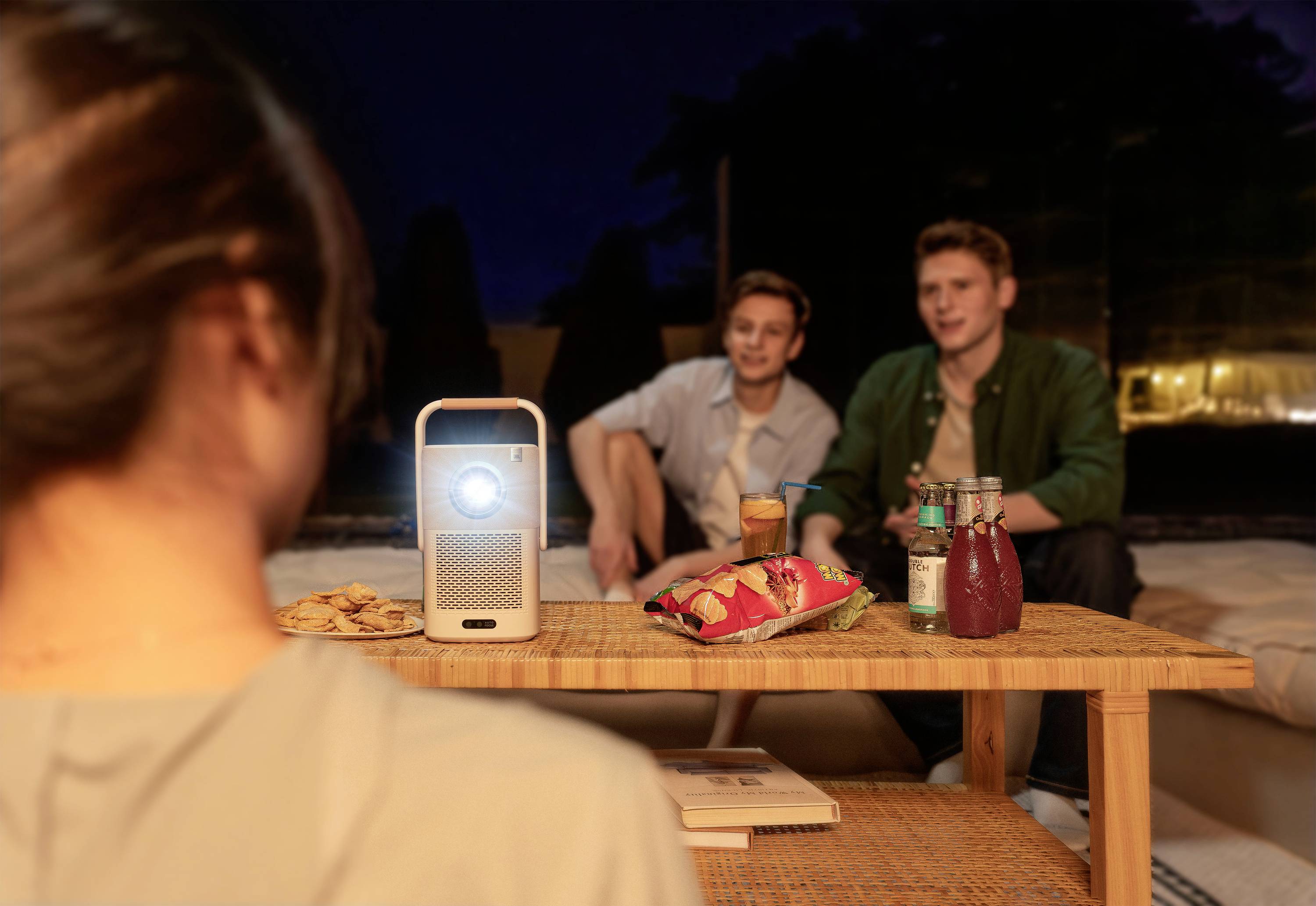 Ein Beamer projiziert ein Bild in einer nächtlichen Outdoor-Szene. Zwei Personen sitzen, Getränke und Snacks stehen auf einem Tisch.