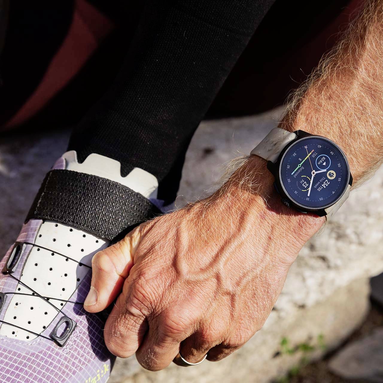Eine Hand schnallt einen Fahrradschuh zu. Am Handgelenk trägt die Person eine Smartwatch, die Uhrzeit und Fitnessdaten anzeigt.