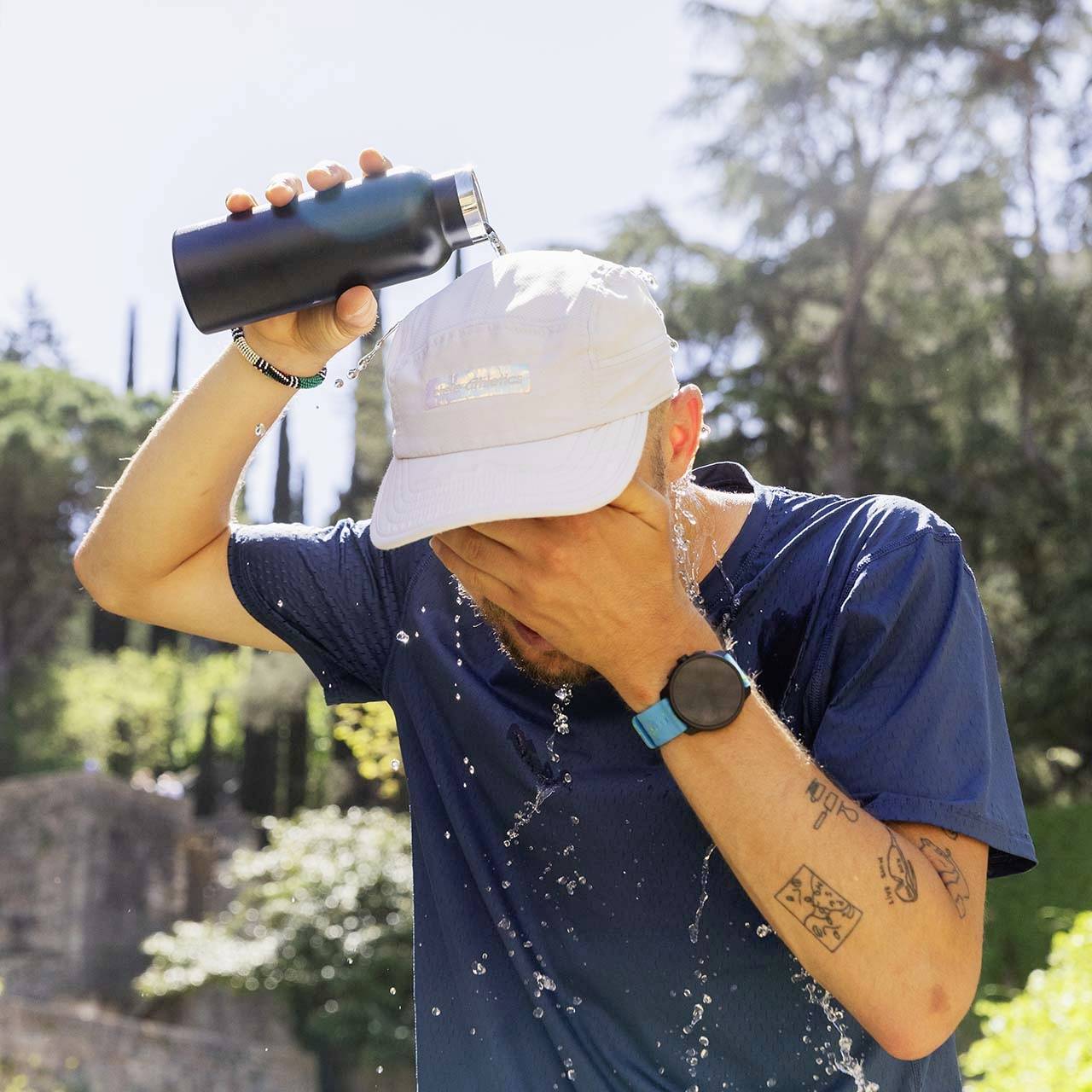 Ein Mann gießt Wasser aus einer Flasche über sein Gesicht, um sich abzukühlen. Im Hintergrund sind Bäume und Pflanzen zu sehen.