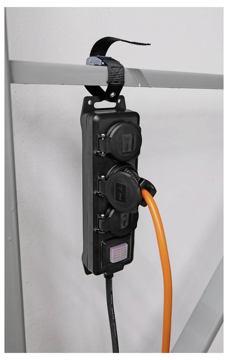 Steckerleiste, die von einer Metallschiene hängt, mit einem schwarzen Kabel und orangefarbenem Kabel, das gegen eine glatte Wand angesteckt ist.