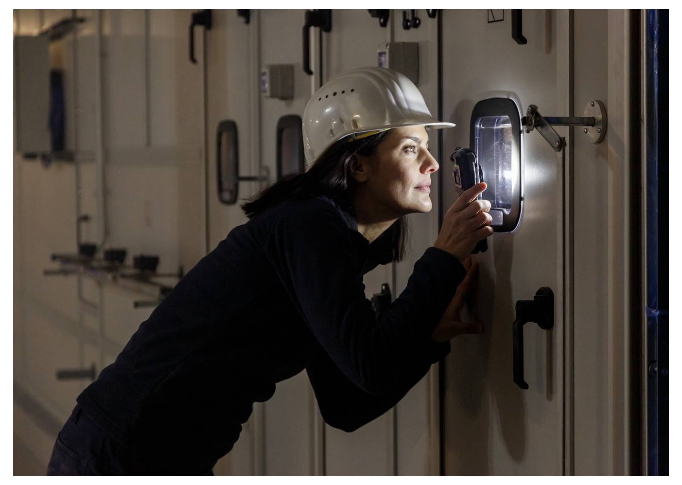 Frau mit Schutzhelm inspiziert ein Türpaneel mit einer Taschenlampe in einem düsteren Industrieflur.