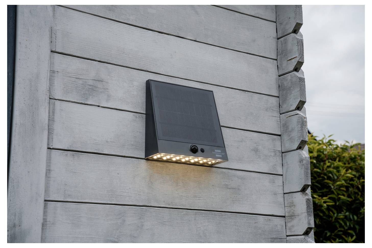 Schwarzes wandmontiertes Solarleuchte mit Bewegungsmelder und leuchtenden LEDs an einer grauen Holzaußenwand.