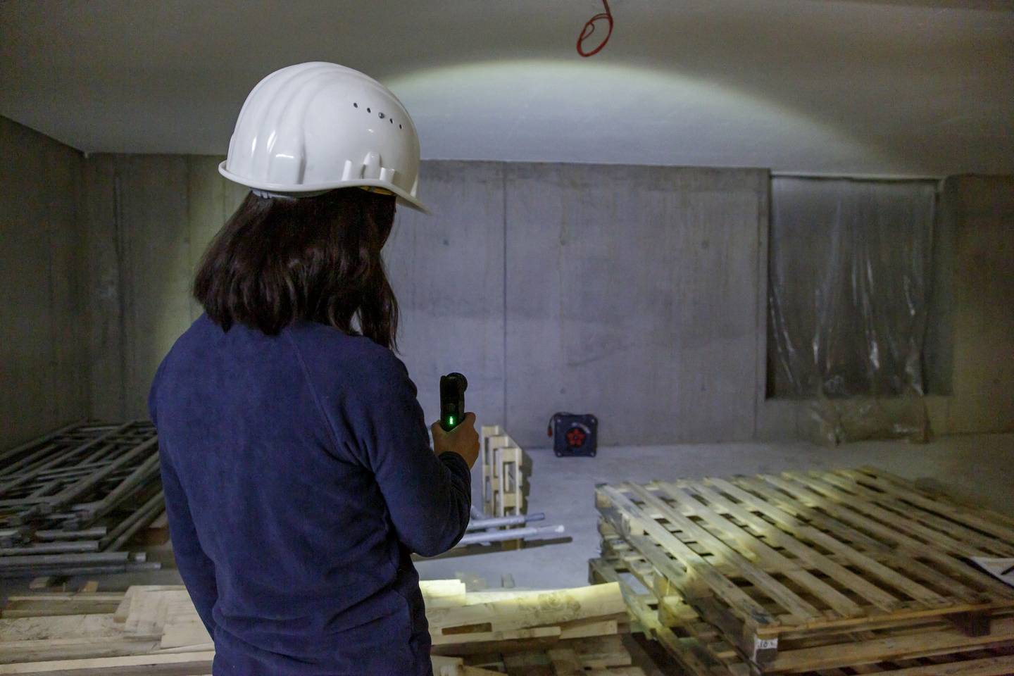 Person mit Helm und Taschenlampe inspiziert Baustelle mit Paletten in einer halbfertigen Betonstruktur.