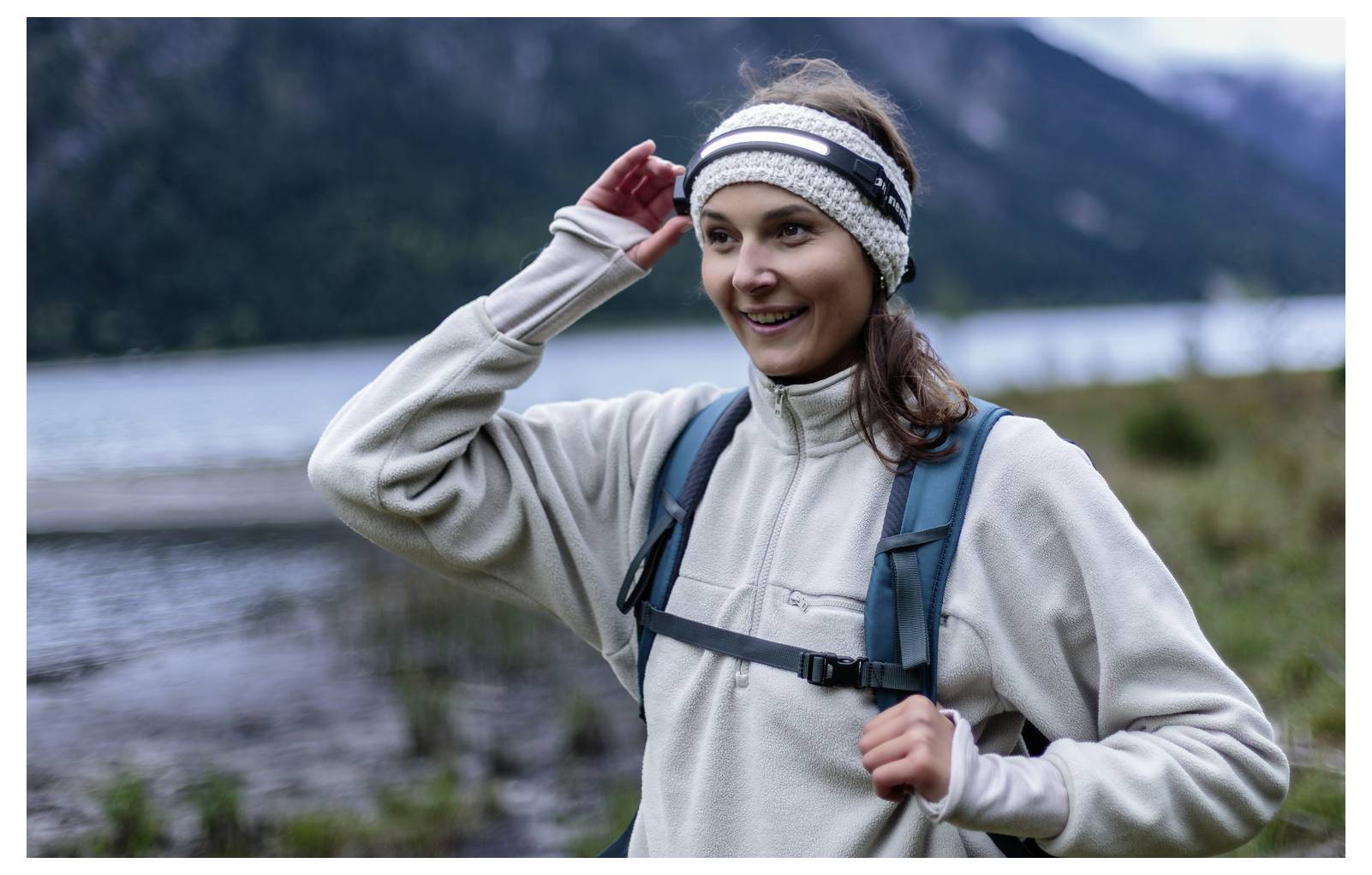 Lächelnde Frau in Outdoor-Kleidung zieht ihren Stirnband zurecht, während sie in der Nähe eines Sees mit Bergen im Hintergrund wandert.