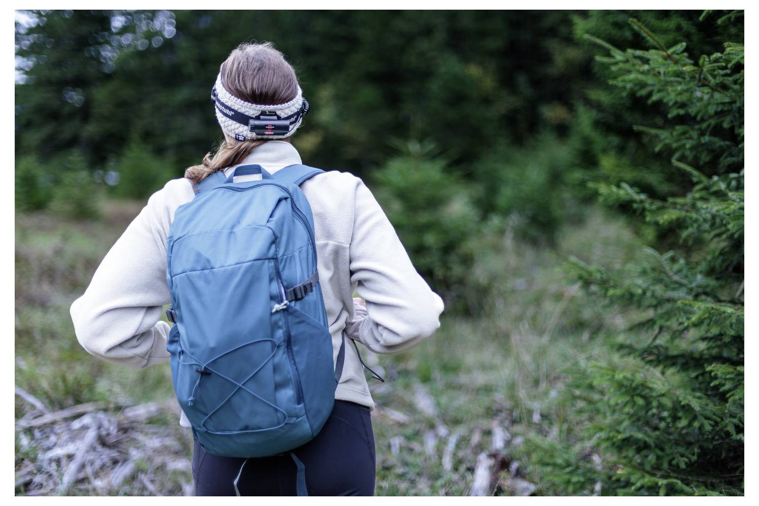 Person mit einem blauen Rucksack steht auf einem Waldpfad, dem Kameramann zugewandt, in Outdoor-Wanderausrüstung.