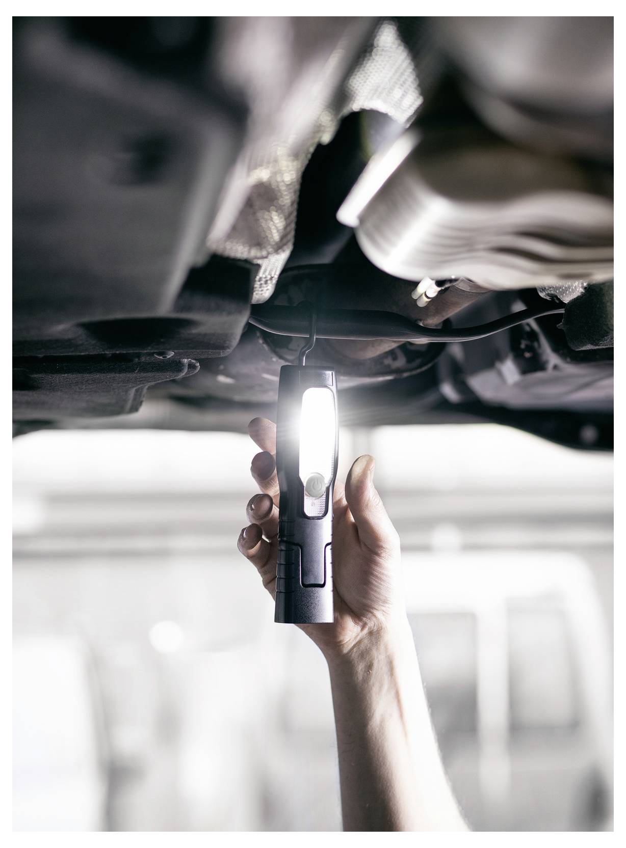 Hand hält eine leuchtende Inspektionsleuchte unter einem Auto und beleuchtet die Unterseite für Fahrzeugwartung oder Reparatur.