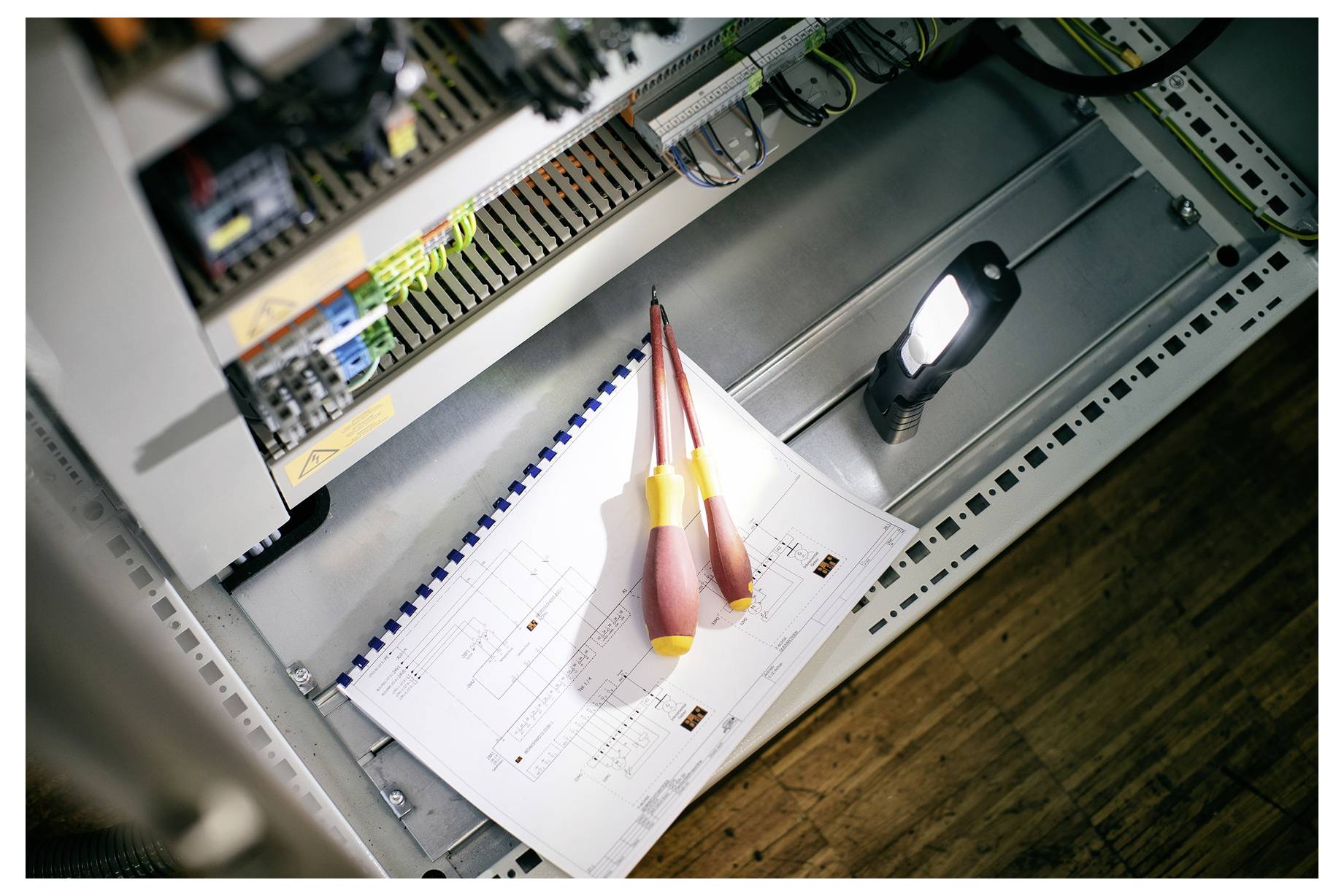 Offenes Elektroschaltfeld mit Leistungsschaltern und Verkabelung über einer Zwischenablage mit technischen Zeichnungen, beleuchtet von einer Taschenlampe.