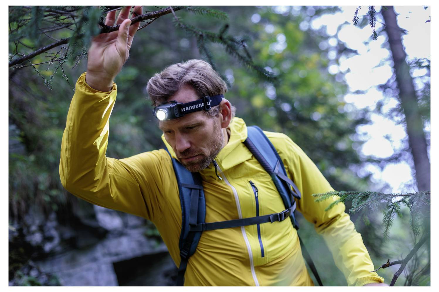 Ein Wanderer in einer gelben Jacke mit einer Stirnlampe hebt einen Arm, während er durch einen Wald geht.