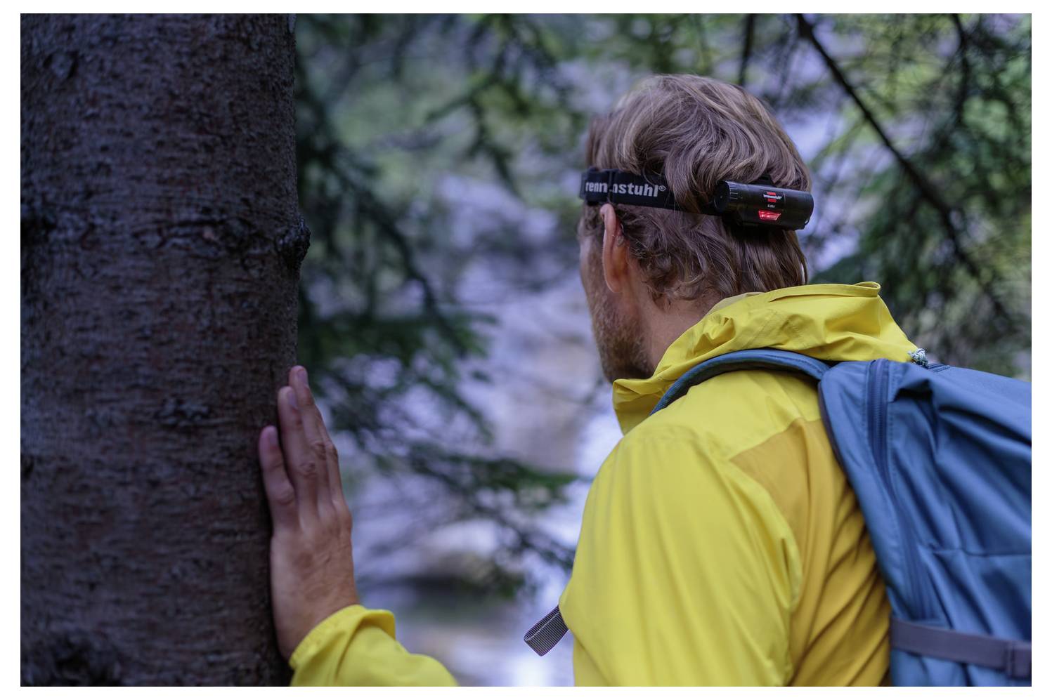 Ein Wanderer in einer gelben Jacke und einem blauen Rucksack drückt eine Hand gegen einen Baum, während er eine Stirnlampe trägt, in einem bewaldeten Gebiet.
