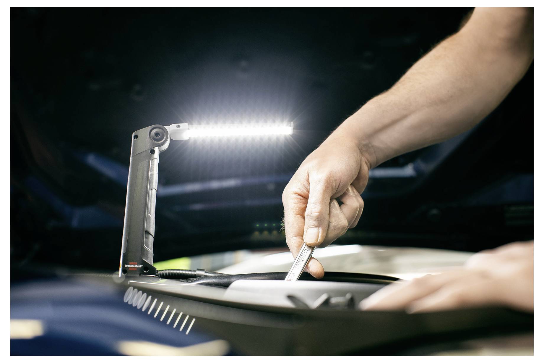Die Hände eines Mechanikers benutzen ein Werkzeug unter einem hellen Arbeitslicht in einem Motorraum während einer Fahzeugreparatur.