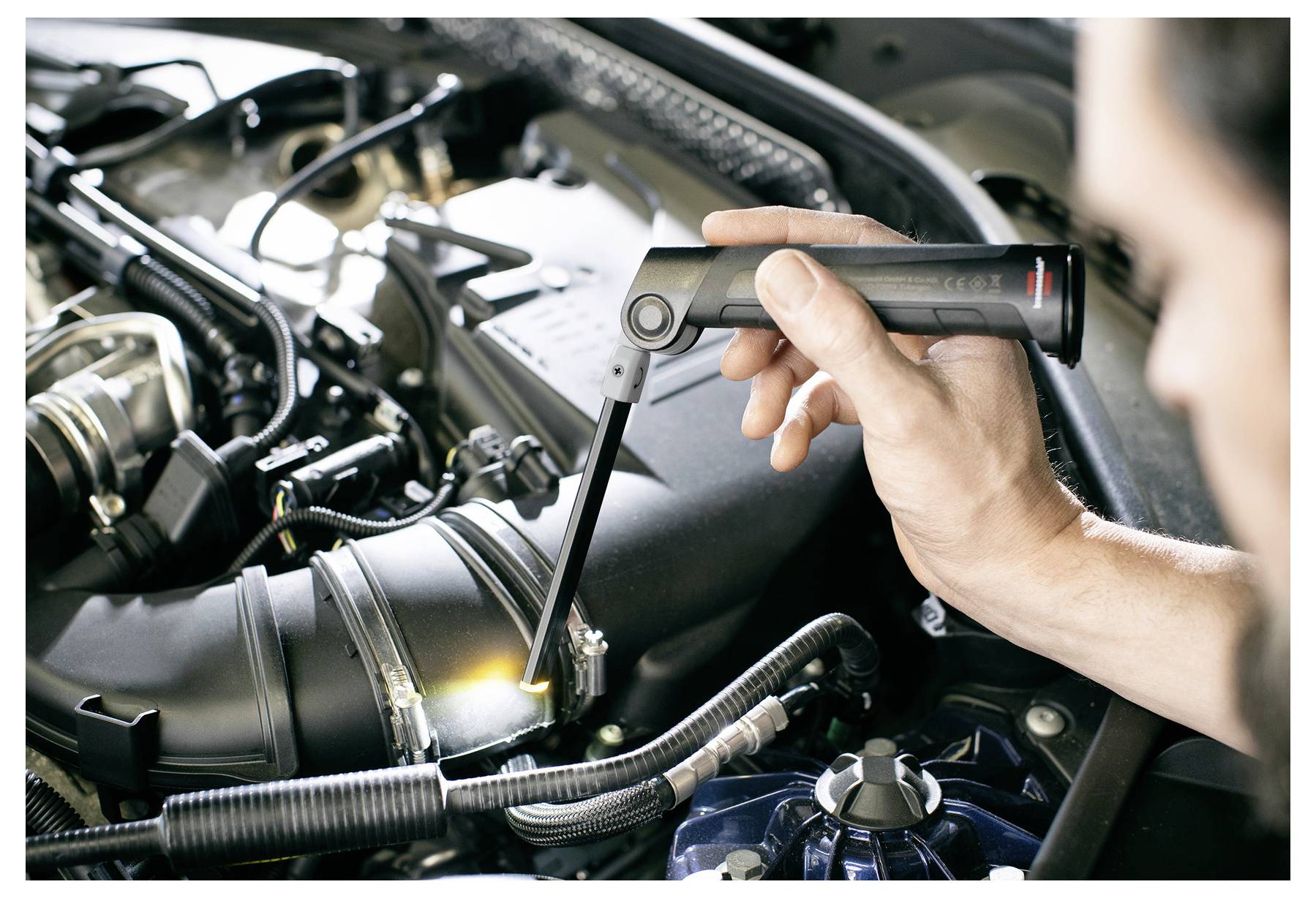 Ein Mechaniker benutzt eine Taschenlampe, um einen Bolzen an einem Automotor unter der Motorhaube zu inspizieren und festzuziehen.