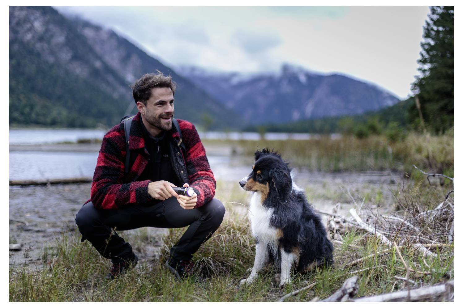 Ein Mann, der neben einem schwarzen, weißen und hellbraun gefärbten Hund in einer grasbewachsenen Seeufer-Berglandschaft hockt.