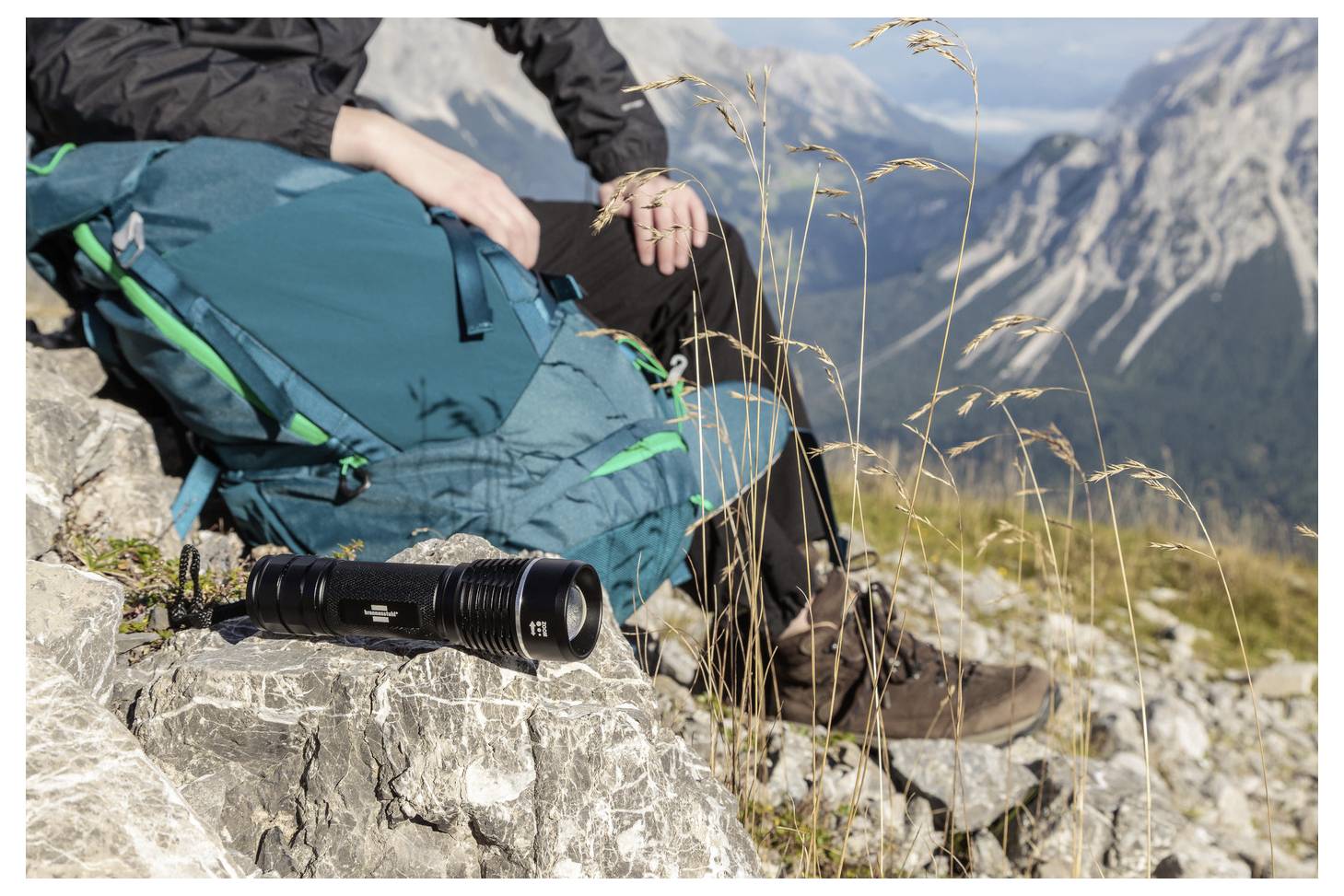 Wanderer ruht sich auf einem felsigen Berghang aus, mit einem blaugrünen Rucksack, Stiefeln und einem Kameraobjektiv im Vordergrund; alpine Gipfel im Hintergrund.