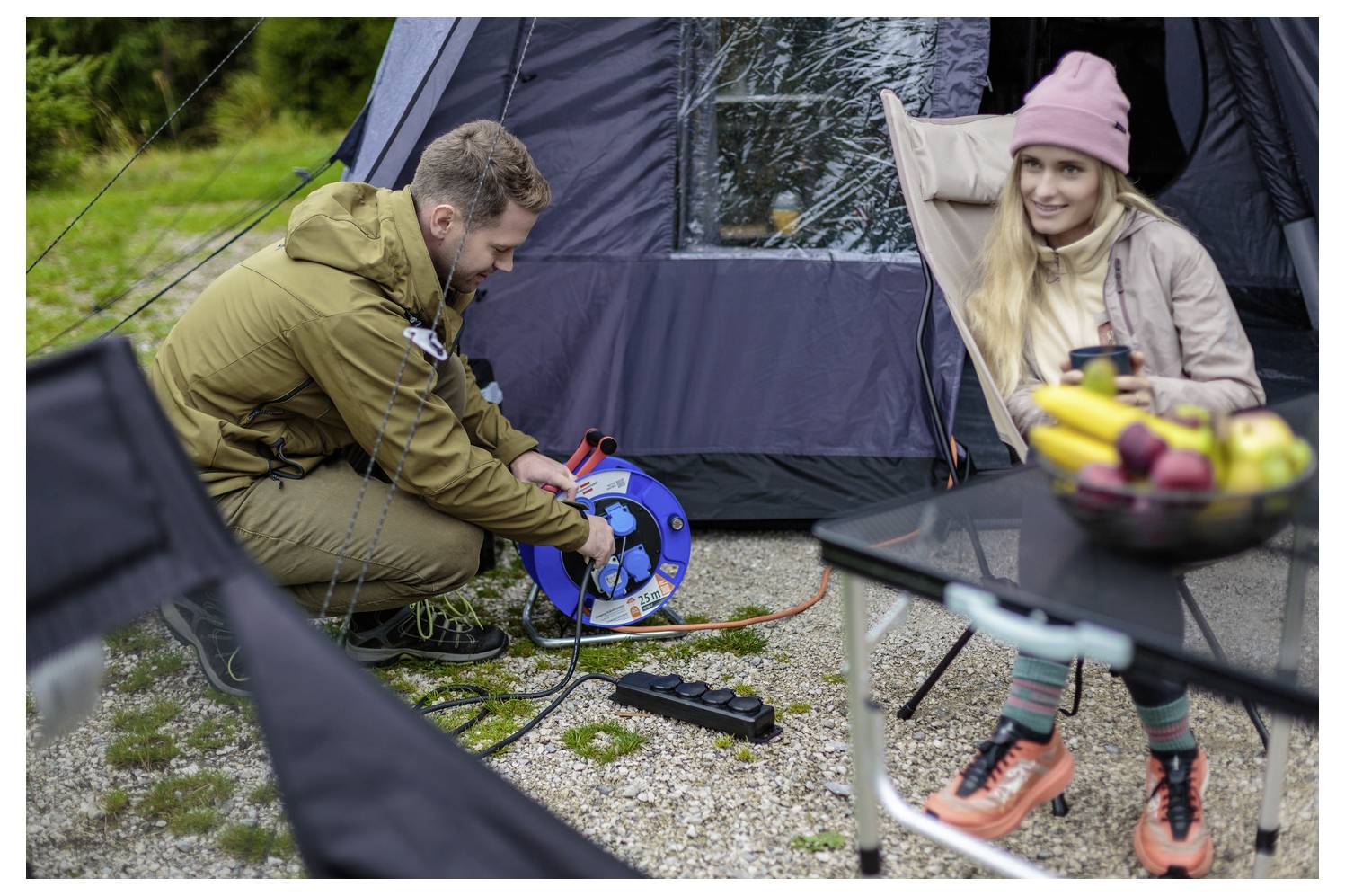 Ein Mann außerhalb eines Zelts wickelt ein Verlängerungskabel auf einer blauen Kabelrolle auf, während eine Frau in der Nähe an einem Campingtisch mit Obst sitzt.