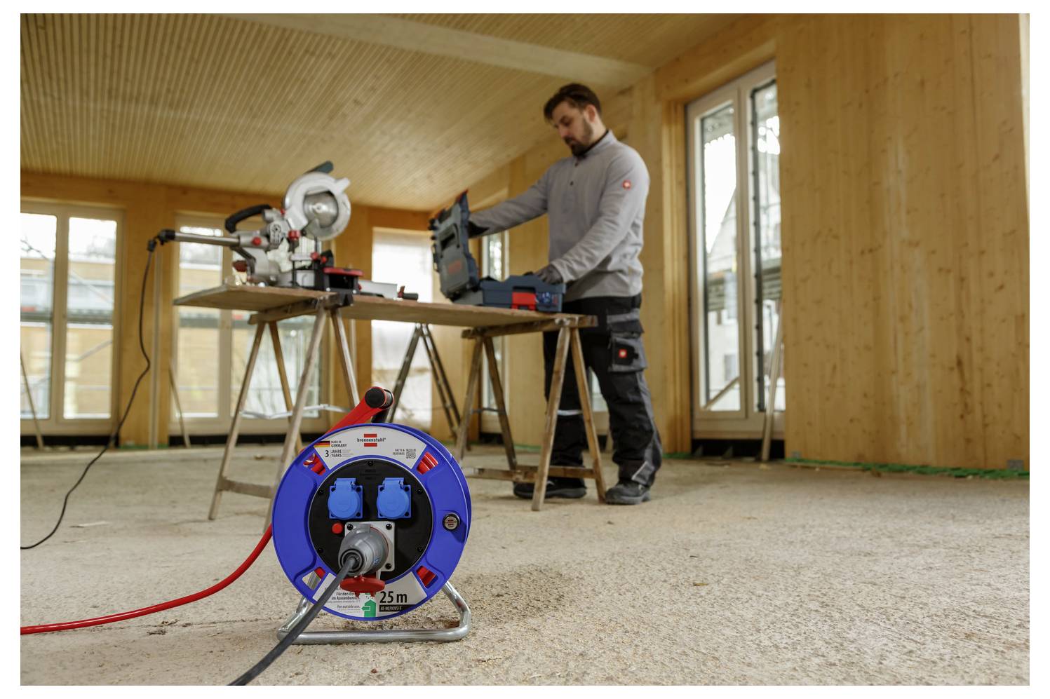 Ein Mann benutzt eine Elektrosäge auf einer Werkbank in einem unvollendeten Raum, mit einer blauen Kabelrolle im Vordergrund.