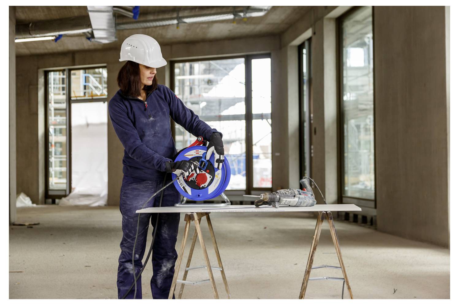 Bauarbeiter in blauem Schutzanzug und weißem Schutzhelm, der eine Elektrosäge auf einem Brett in einem unvollendeten Gebäudeinneren benutzt.