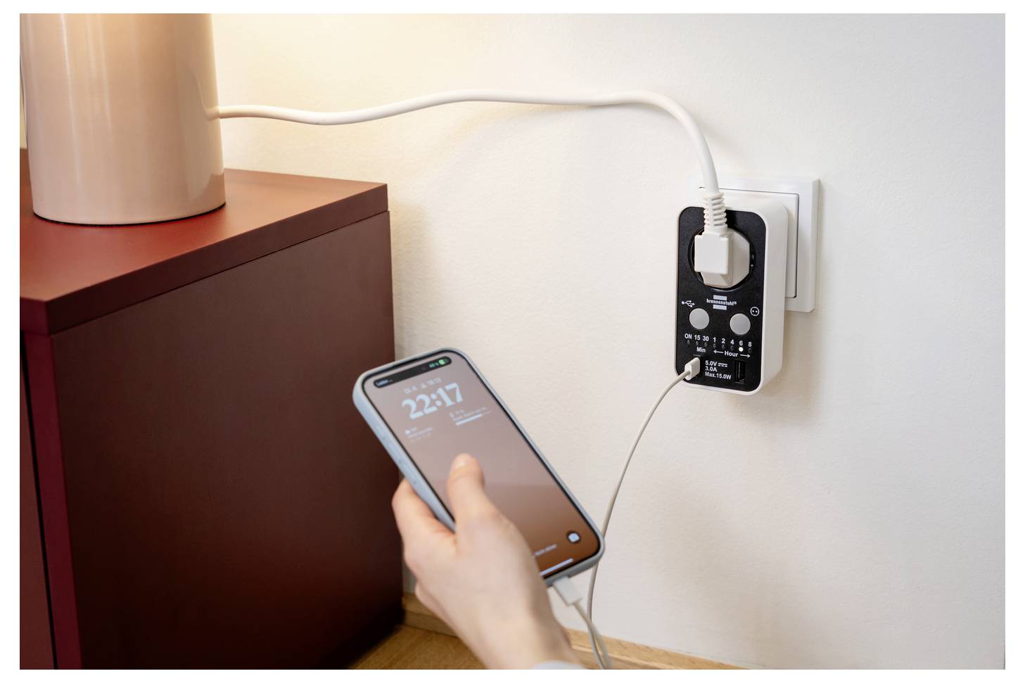 Eine Hand hält ein Smartphone, das an einen Wandladegerät angeschlossen ist, neben einem Schrank, wobei das Telefon über einen Steckdosenadapter aufgeladen wird.