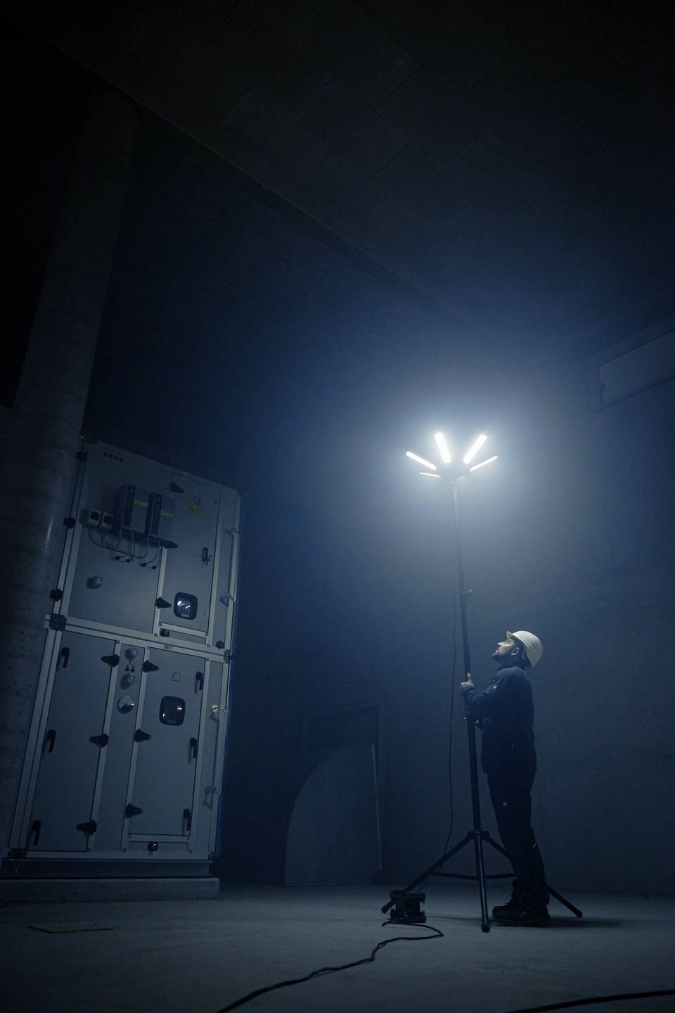Eine Person in Sicherheitskleidung justiert eine helle Leuchte auf einem Stativ in einem dunklen, industriellen Raum.