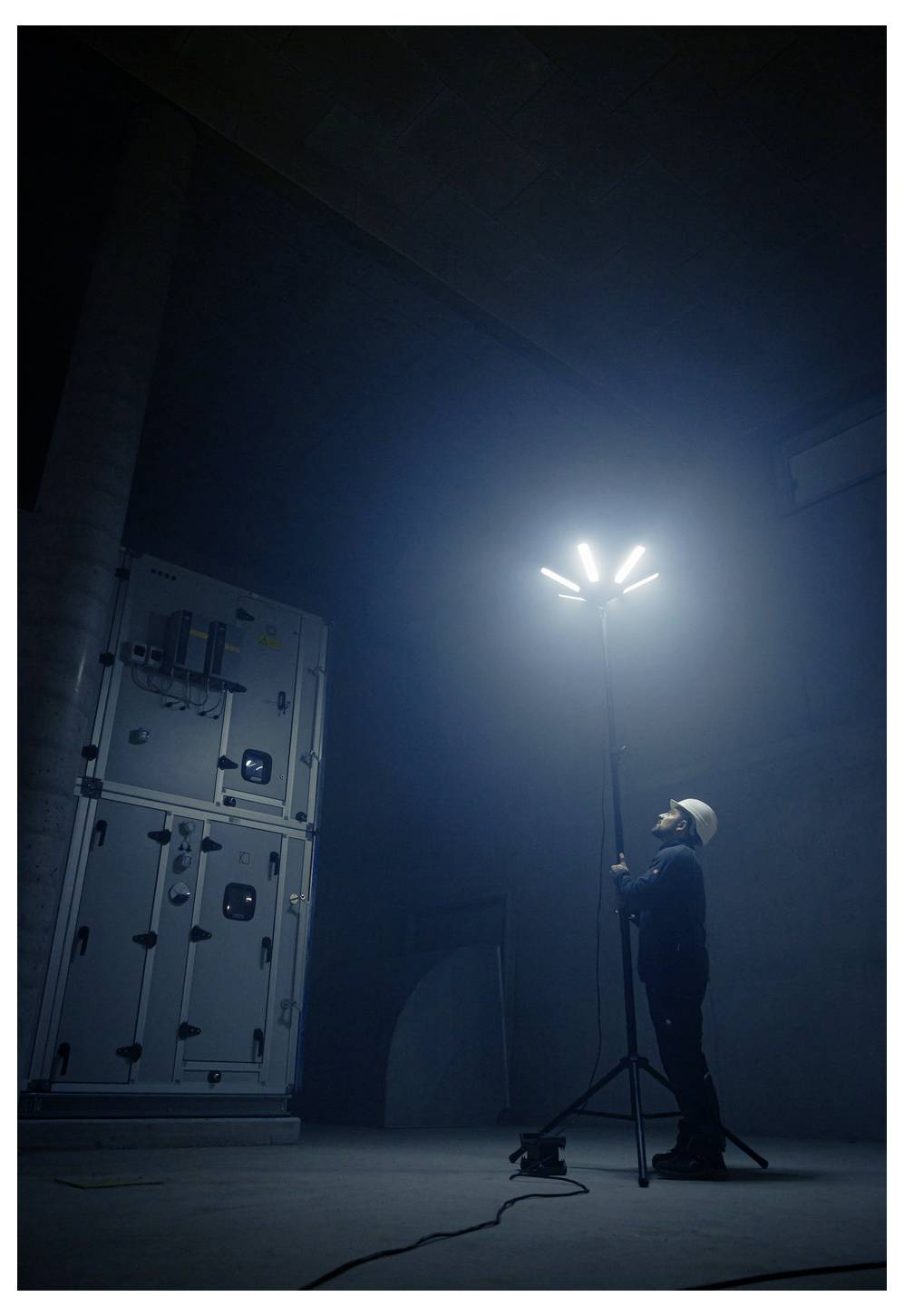 Arbeiter mit Helm stellt ein hohes Stativarbeitslicht in einem dunklen Industrieraum neben großen Geräten ein.
