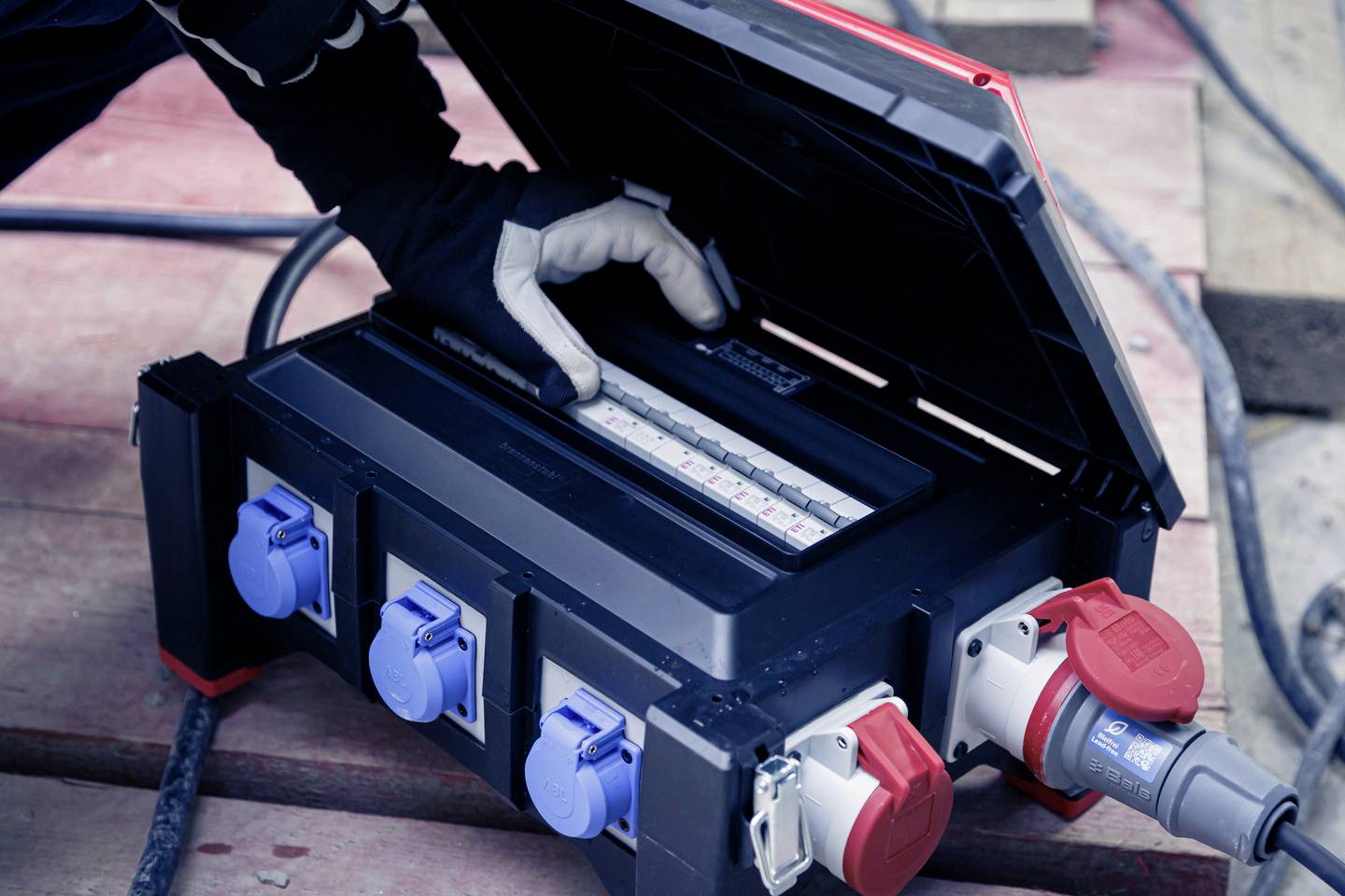 Eine Person in Arbeitshandschuhen öffnet einen Verteilerkasten auf einer Baustelle, überprüft oder justiert Schaltgeräte und Anschlüsse.