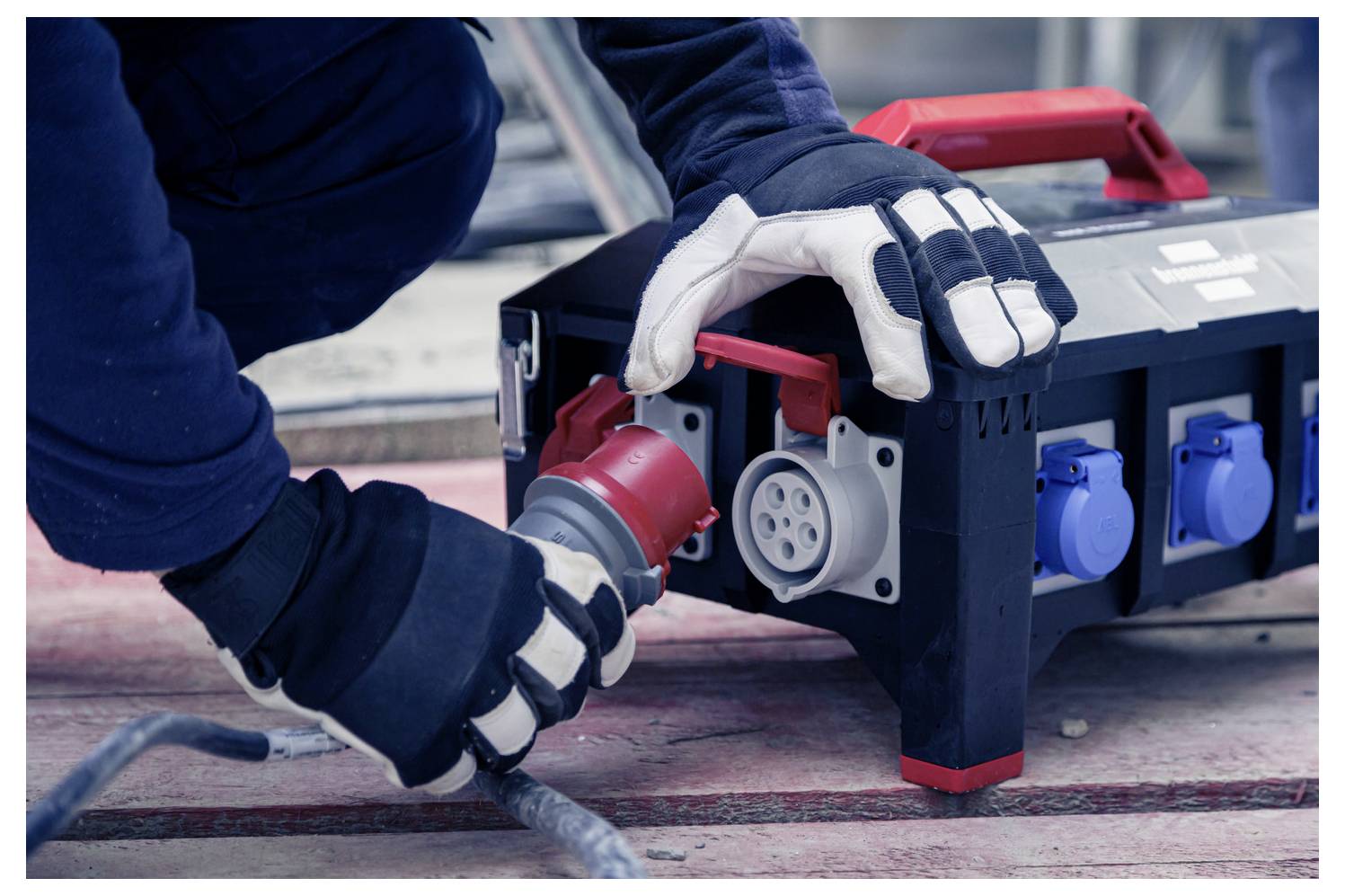 Ein Arbeiter mit Handschuhen steckt einen roten Industrie-Stromstecker in eine tragbare Elektroverteilungsbox auf dem Boden.