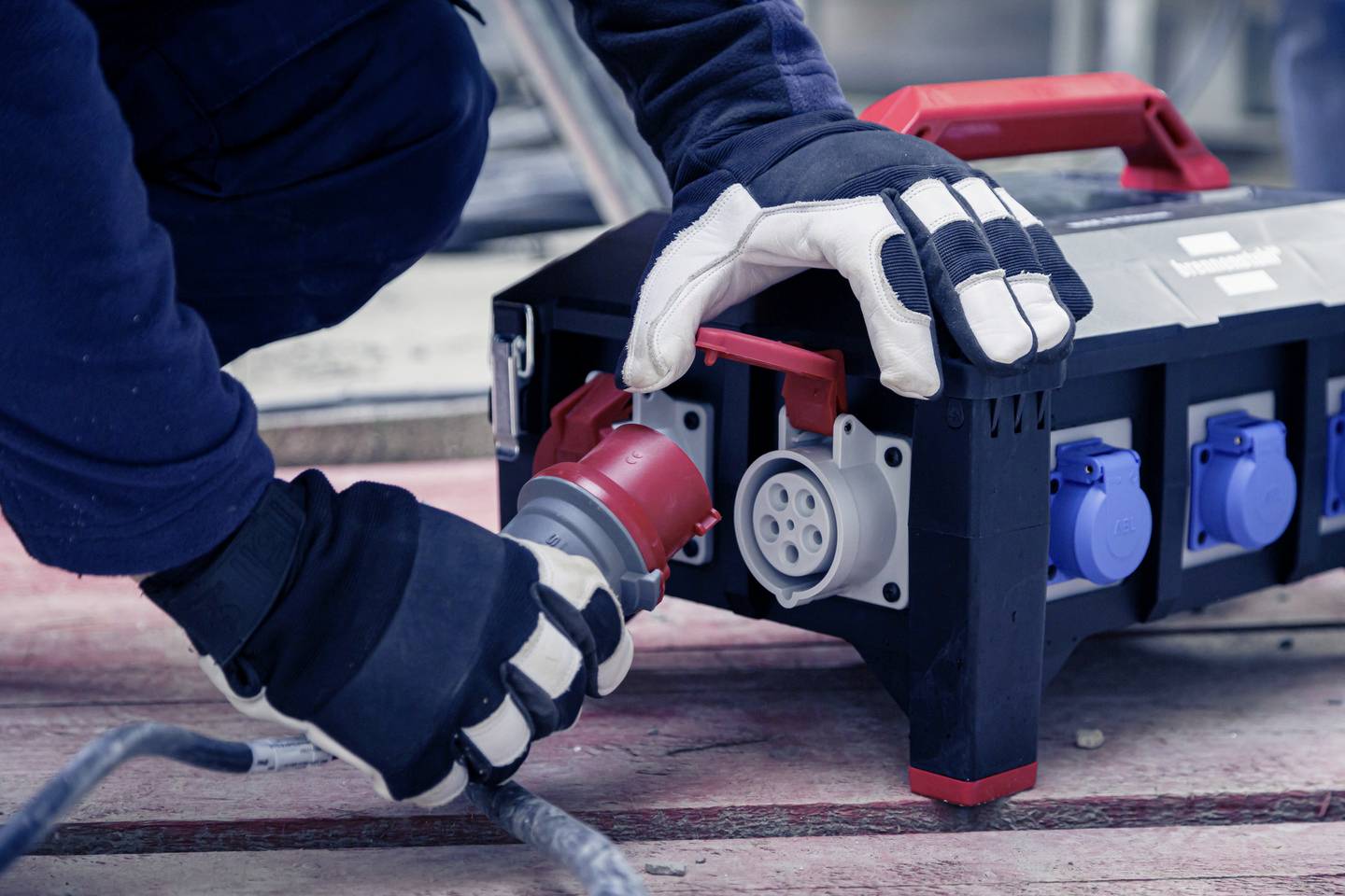 Eine Person steckt einen großen Stecker in eine industrielle Stromverteilereinheit ein. Es wird auf einer Baustelle gearbeitet.