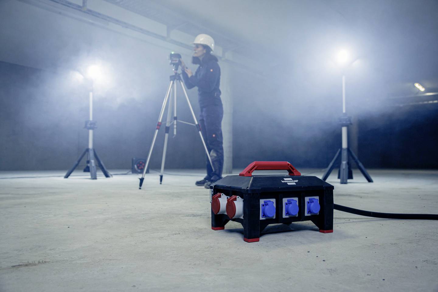 Ein Techniker in Schutzkleidung arbeitet in einem nebligen Raum, daneben ein Verteilerkasten mit Steckdosen, im Hintergrund starke Scheinwerfer.