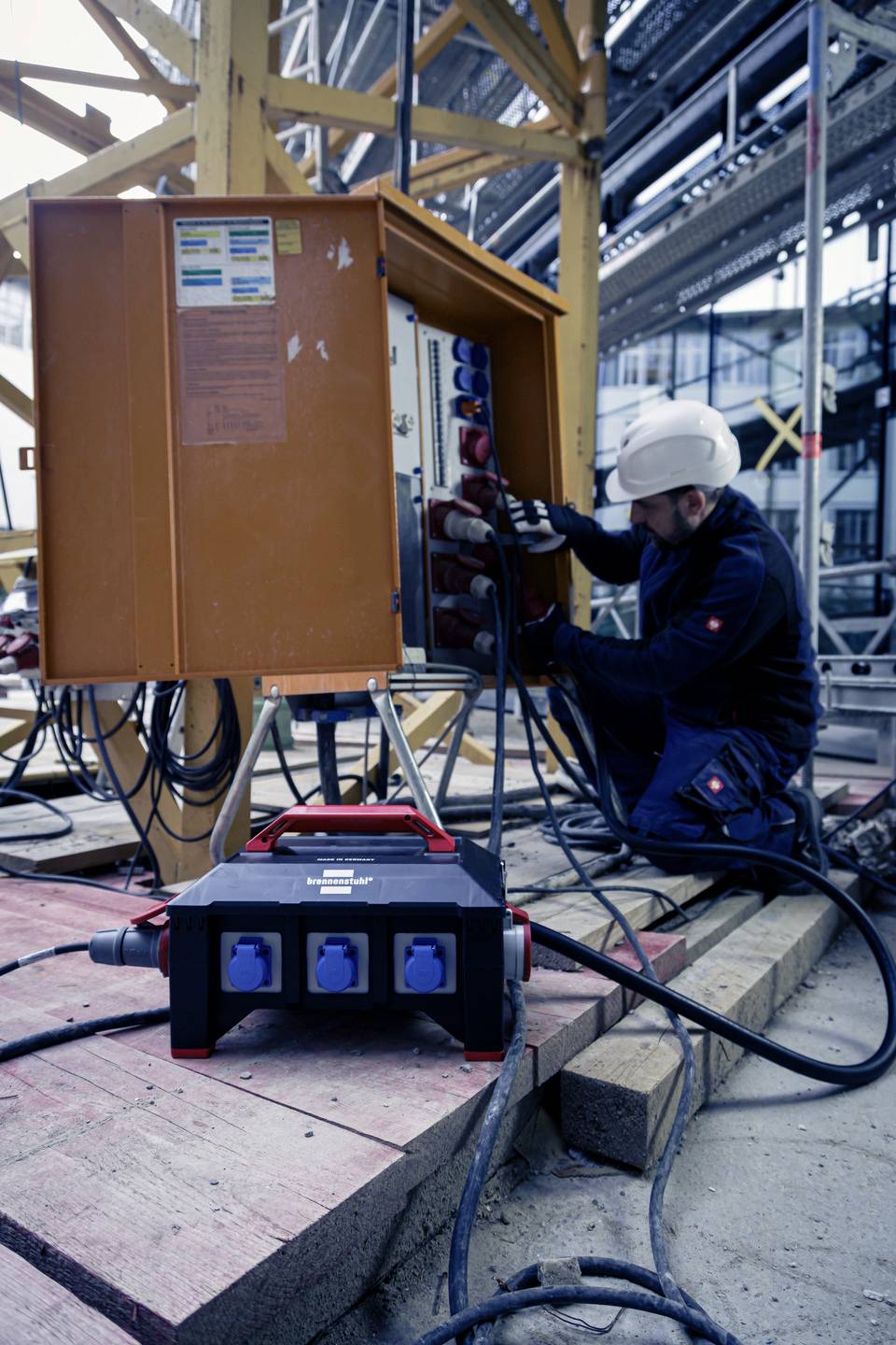 Techniker arbeitet an einem Baustromverteilerkasten auf einer Baustelle, trägt Schutzhelm und Arbeitskleidung.