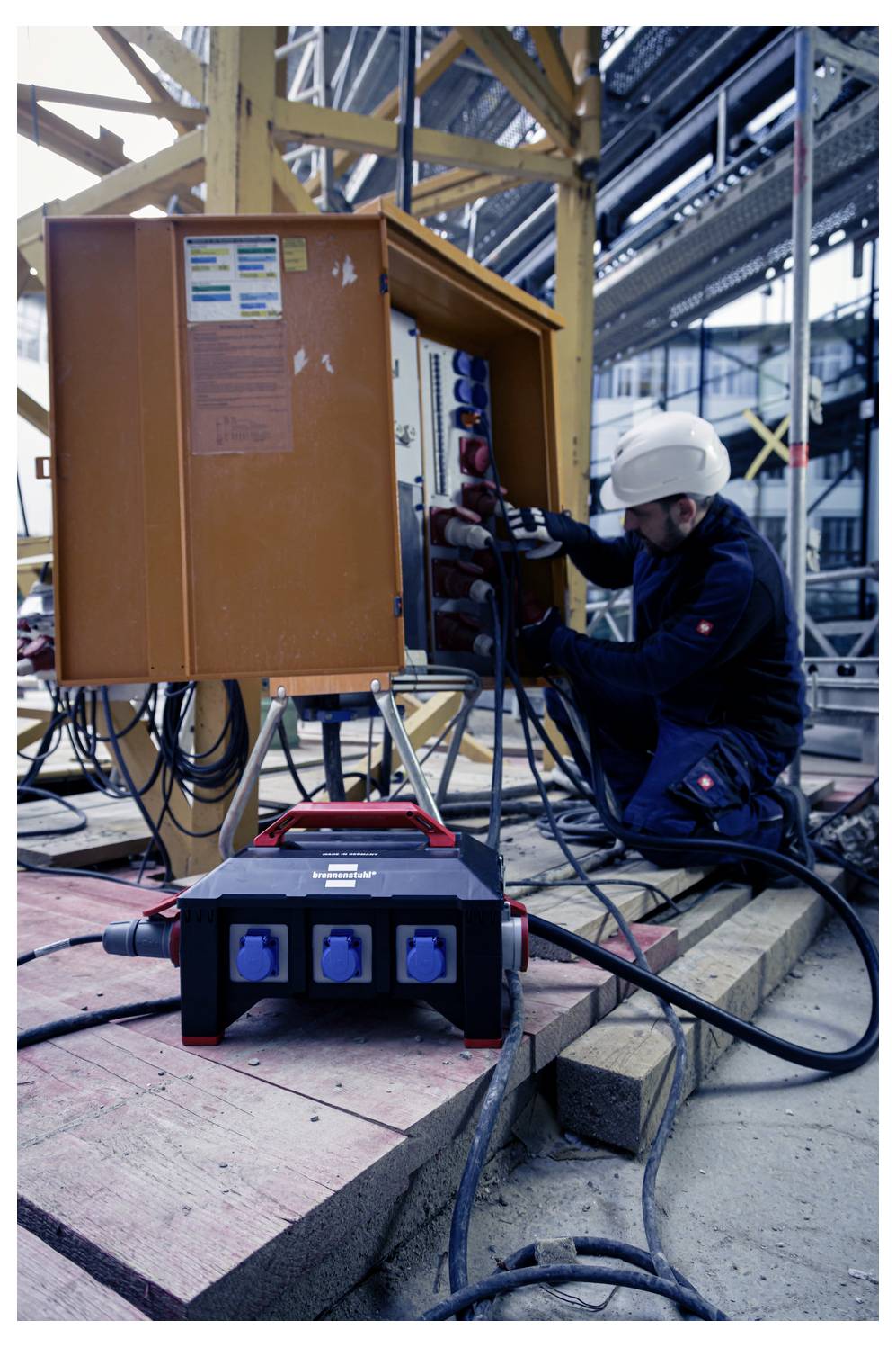 Bauarbeiter in Schutzhelm verkabelt einen elektrischen Schaltschrank auf einer Baustelle, mit einer tragbaren Stromverteilungsbox im Vordergrund.