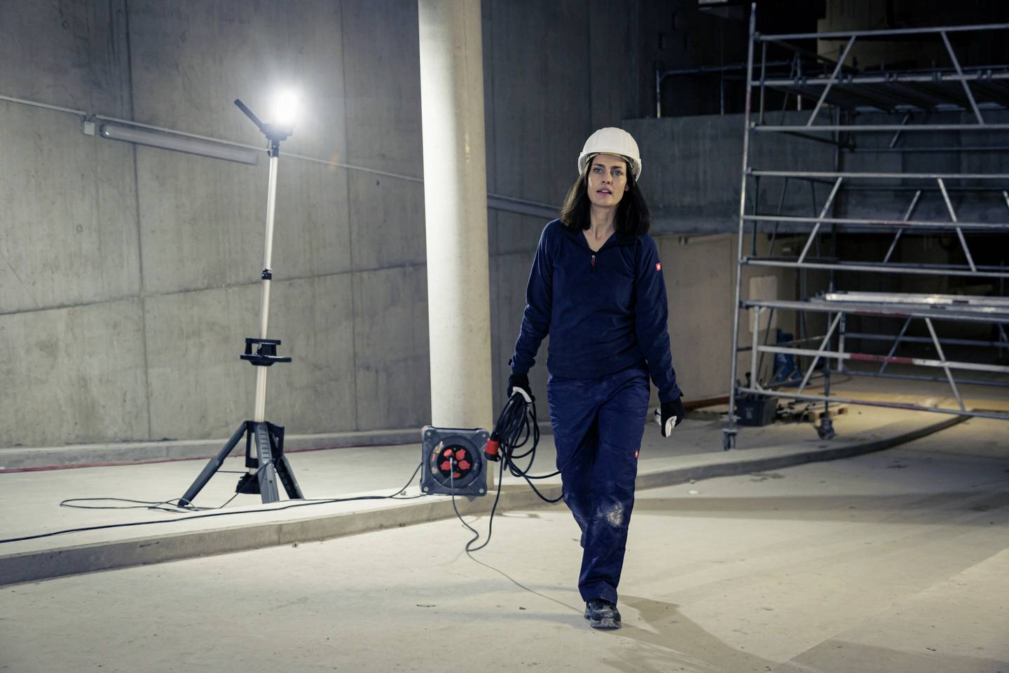 Eine Frau in Arbeitskleidung und Schutzhelm läuft in einem unfertigen Gebäude. Im Hintergrund steht ein Baustrahler und ein Gerüst.