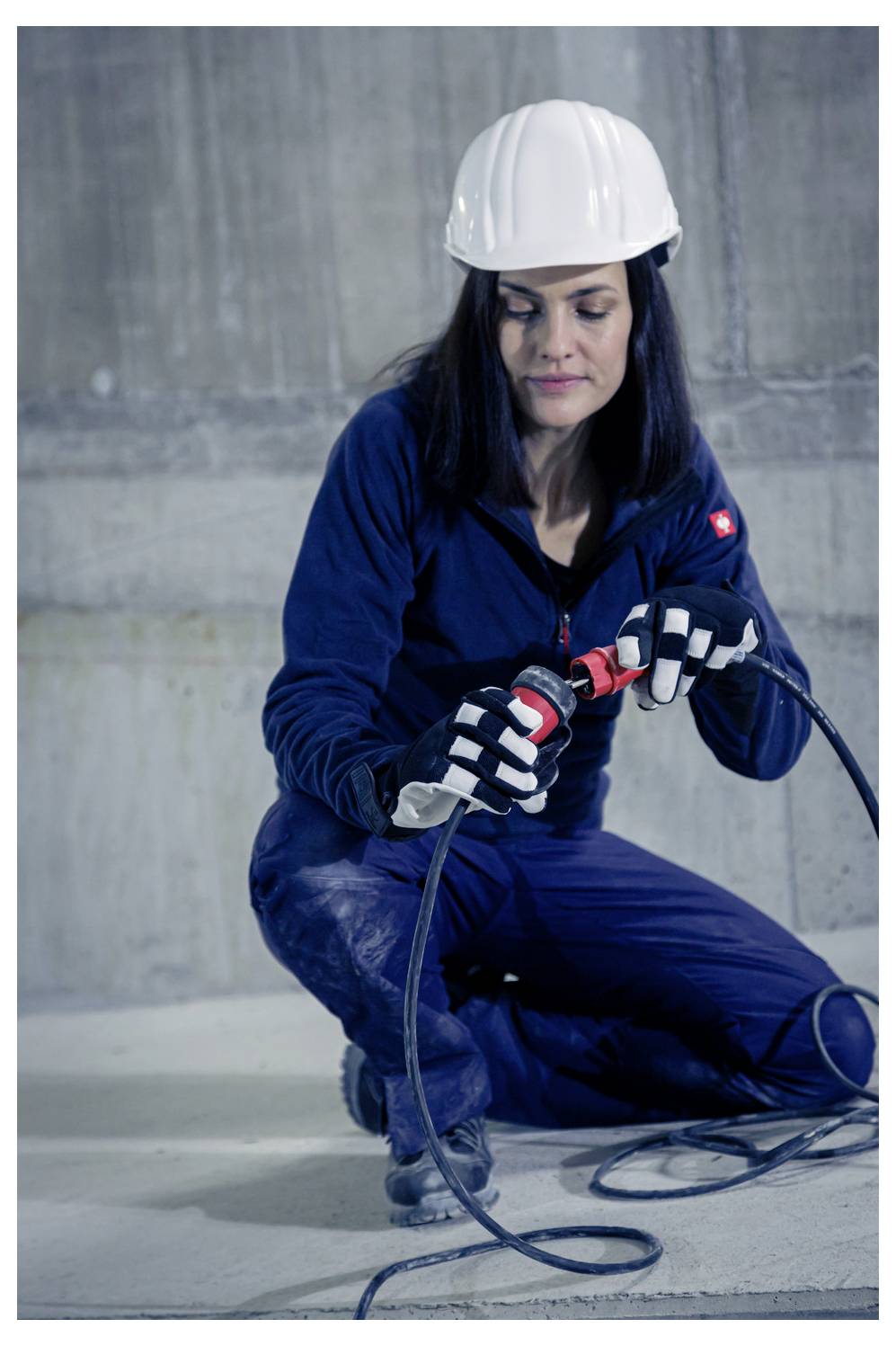 Weibliche Bauarbeiterin, die auf einem Betonboden kniet, ein elektrisches Werkzeug mit Kabel verwendet und einen Schutzhelm, Handschuhe und einen blauen Arbeitsanzug trägt.