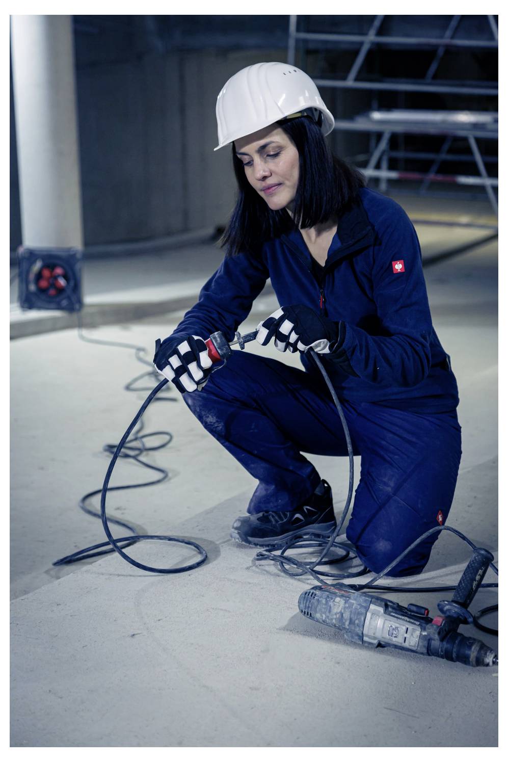 Weibliche Bauarbeiterin mit Schutzhelm, die auf dem Boden kniet und ein Elektrowerkzeug verwendet, mit Kabeln und Ausrüstung um sie herum auf einer Baustelle.