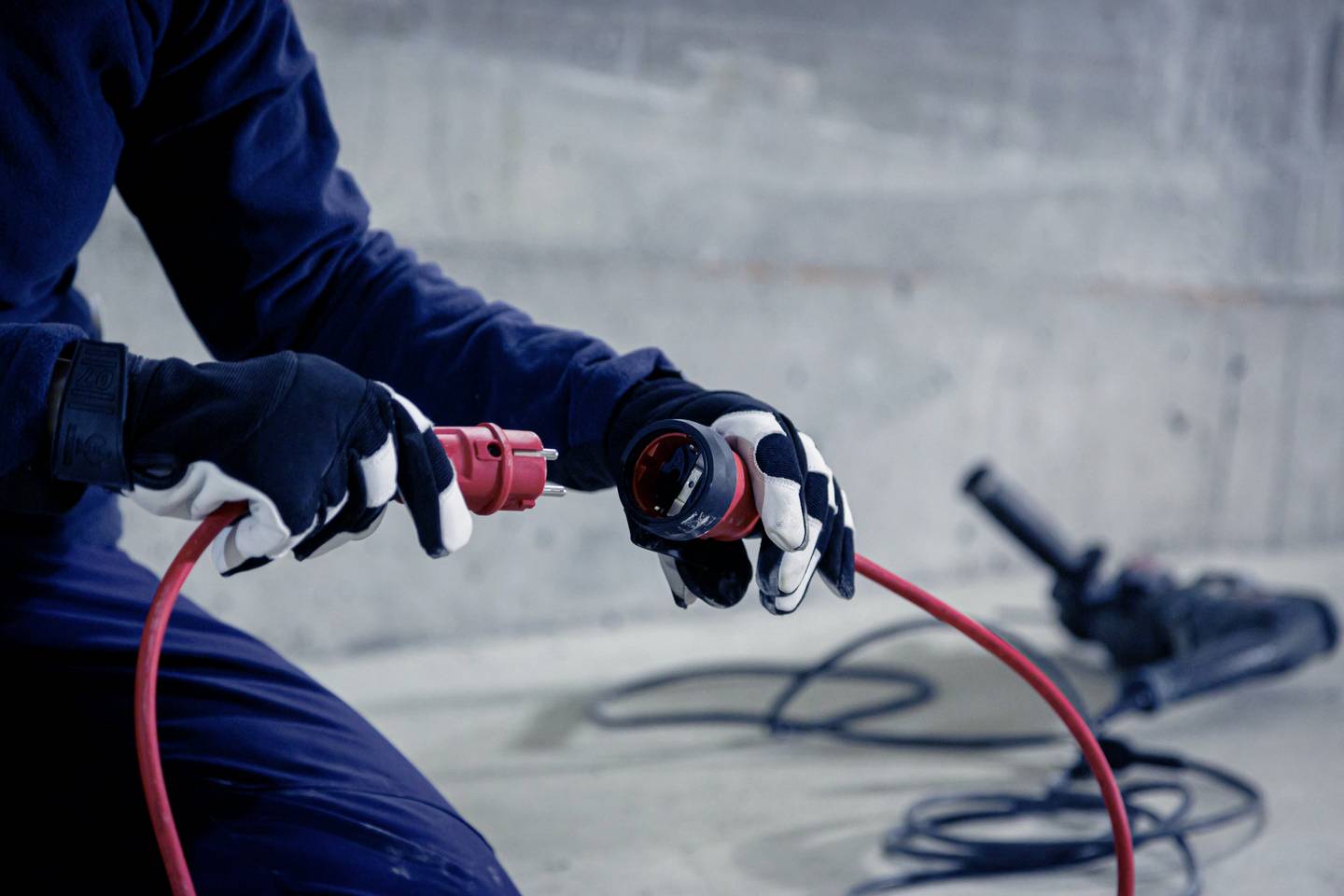 Eine Person in Arbeitshandschuhen schließt ein rotes Stromkabel in einer industriellen Umgebung an. Im Hintergrund ein Werkzeug.