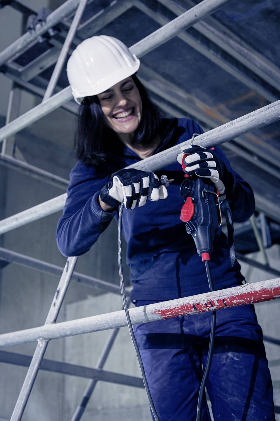 Eine Person in Arbeitskleidung und Schutzhelm benutzt einen Bohrer auf einem Baugerüst. Sie lächelt und konzentriert sich auf die Arbeit.