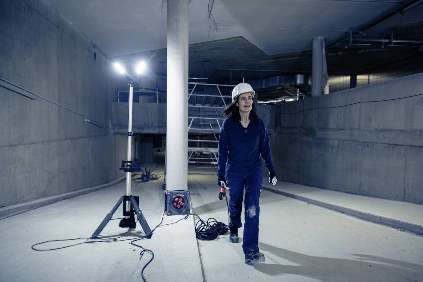 Eine Person in Schutzkleidung und Helm geht durch eine Baustelle mit Betonwänden, beleuchtet von einem Baulicht. Im Hintergrund sind Baugerüste.