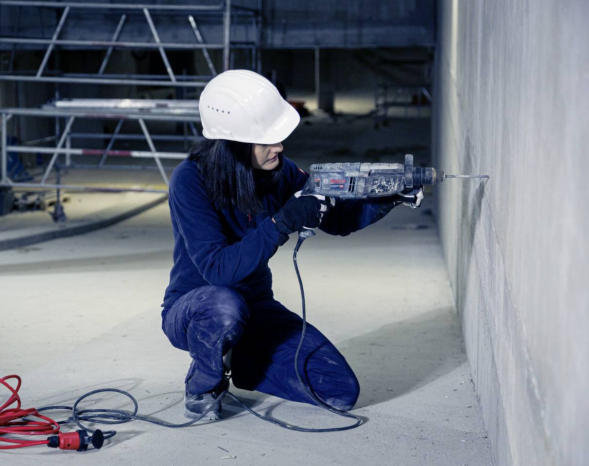 Eine Person trägt Schutzkleidung und verwendet eine Bohrmaschine, um in eine Betonwand zu bohren. Im Hintergrund ist ein Gerüst erkennbar.