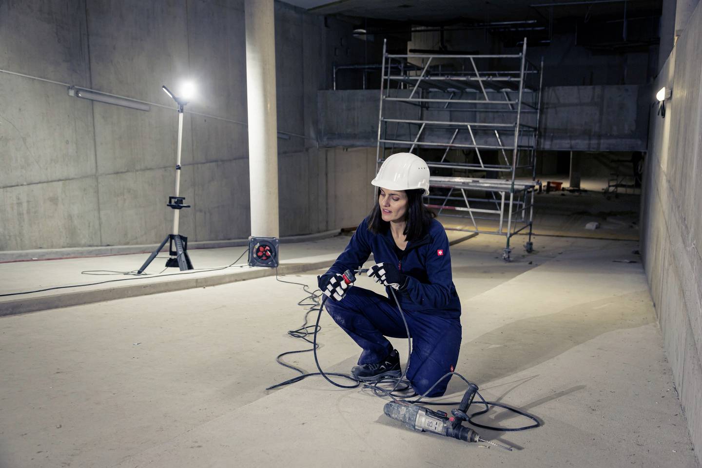 Eine Frau in Arbeitskleidung und Schutzhelm kniet auf einer Baustelle, hält ein Kabel und einen Bohrer. Im Hintergrund steht ein Gerüst.