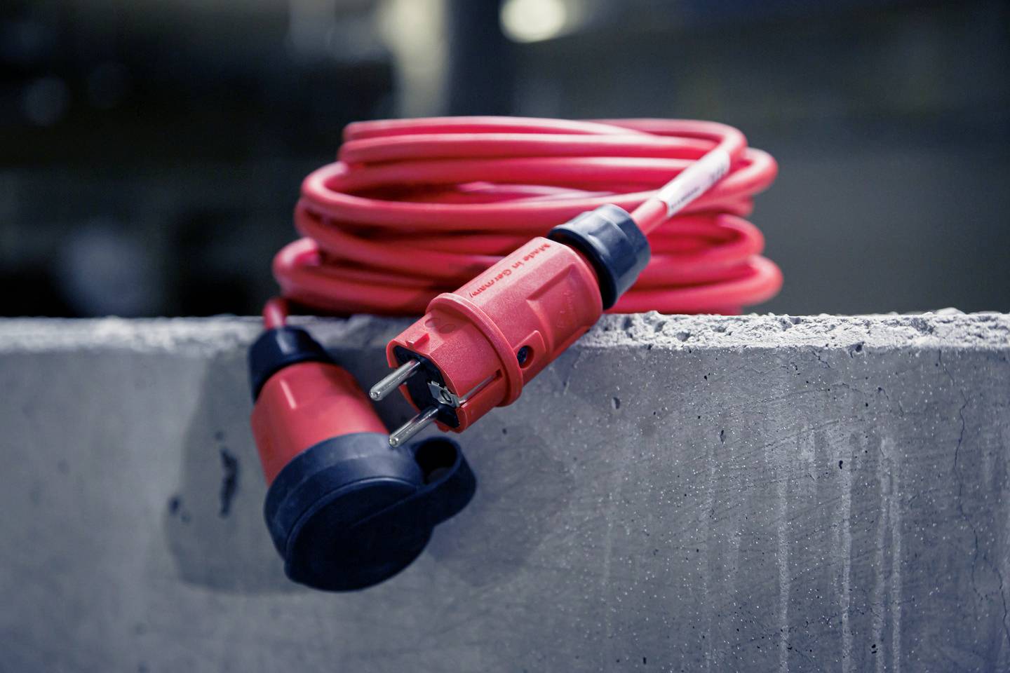 Rotes Verlängerungskabel mit Stecker und Kupplung liegt auf Betonfläche. Kabel ist aufgerollt, Stecker zeigt nach links.