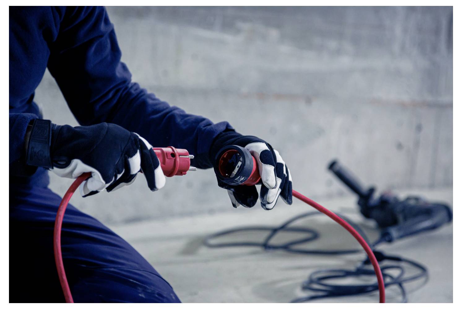 Ein handschuhgetragener Arbeiter verbindet ein rotes Stromkabel mit einem tragbaren Stecker, wobei aufgerollte Kabel auf einem Betonboden liegen.