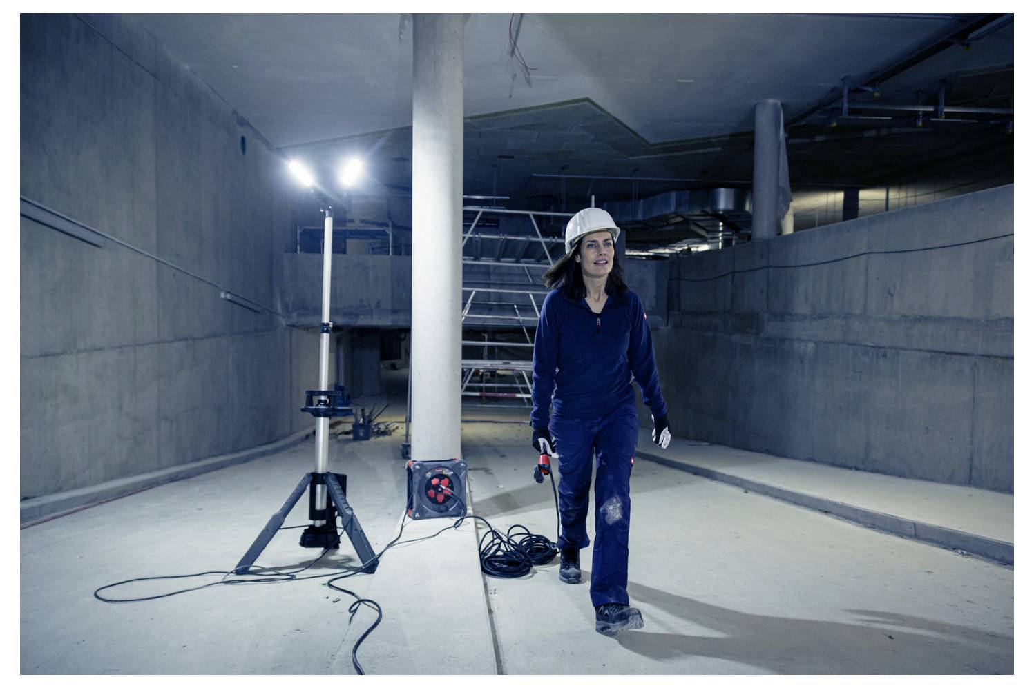 Arbeiterin mit Schutzhelm geht durch einen unvollendeten Betontunnel, mit Baumaschinen und hellen Arbeitsleuchten hinter ihr.