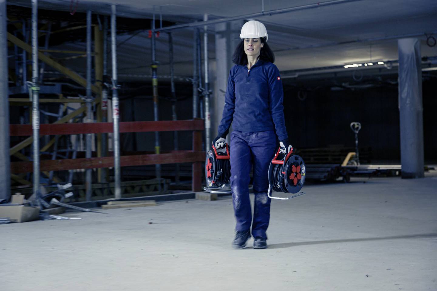 Eine Person auf einer Baustelle trägt zwei Kabeltrommeln. Sie trägt einen Schutzhelm und Arbeitskleidung.