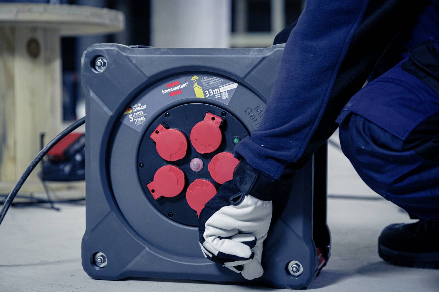 Ein Arbeiter in Schutzkleidung überprüft ein Verlängerungskabel, das auf einer Kabeltrommel aufgewickelt ist, in einer industriellen Umgebung.