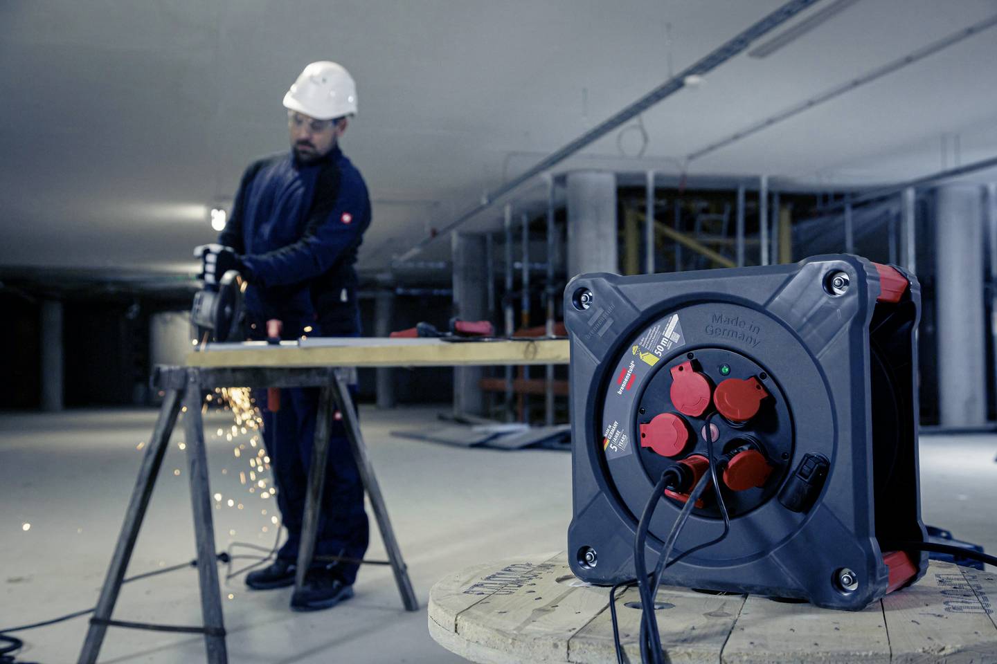 Ein Arbeiter mit Schutzhelm benutzt ein Schleifgerät, Funken fliegen. Im Vordergrund steht eine Kabeltrommel.