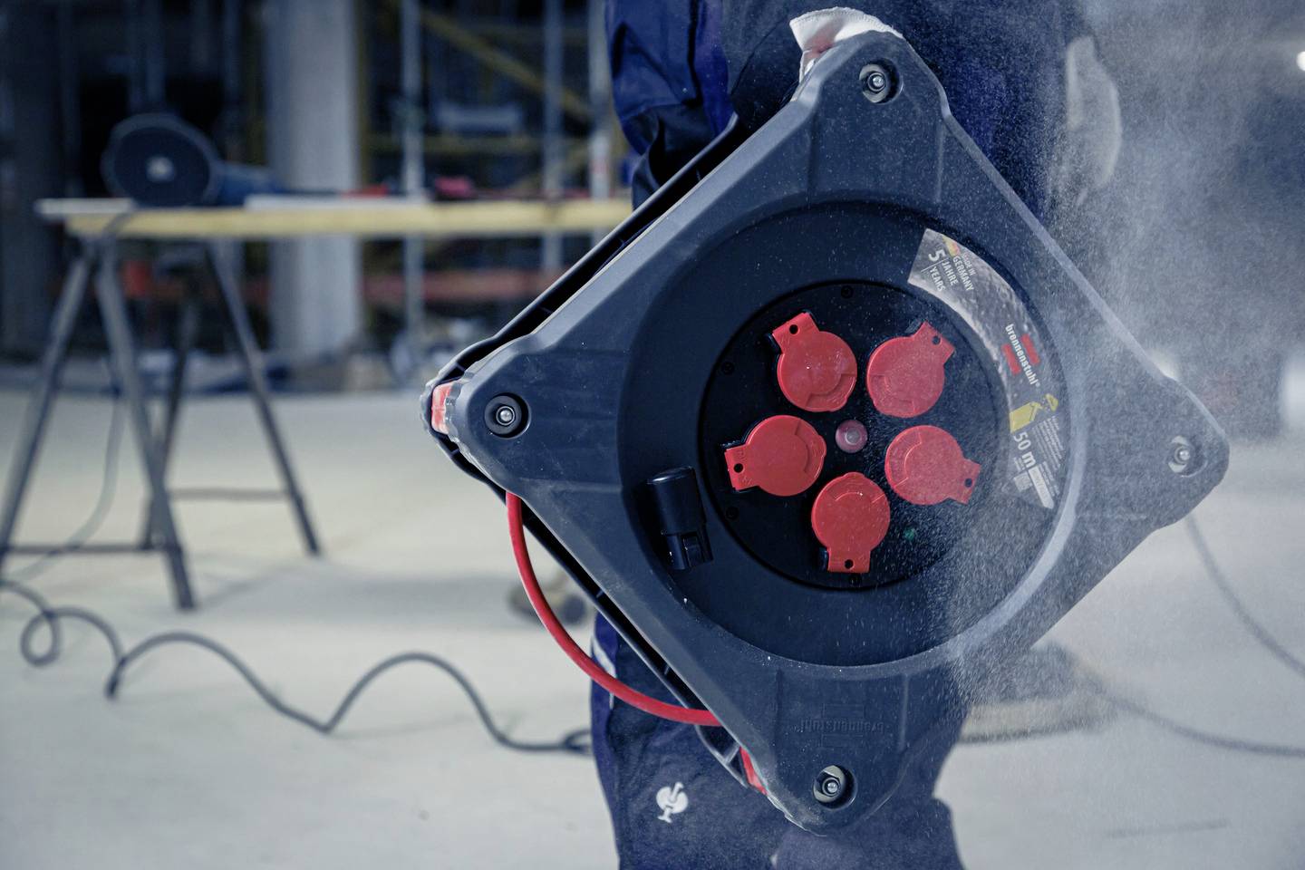 Eine Person hält eine Kabeltrommel mit mehreren Anschlüssen in einer Werkstattumgebung. Im Hintergrund sind Werkzeuge sichtbar.