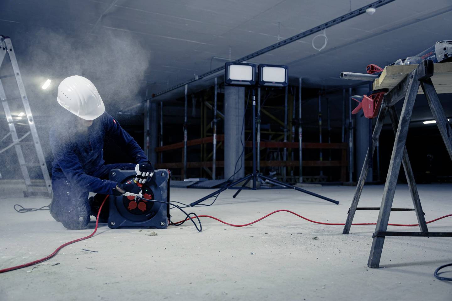 Arbeiter in Schutzkleidung bedient ein Kabeltrommelgerät auf einer Baustelle. Es herrscht rauchige Atmosphäre und Baustellenbeleuchtung.